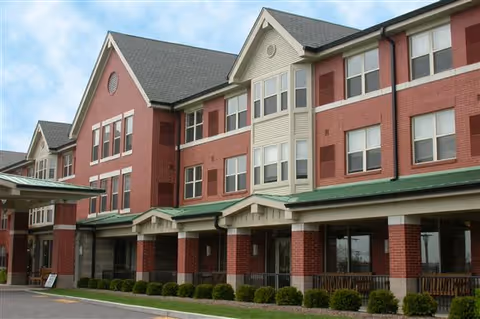 Exterior view of McCormack House-Forest Park Southeast, a multi-story senior living facility with red brick and beige siding, multiple windows, and a covered entrance with green roofing. There are small bushes and a paved driveway in front of the building under a partly cloudy sky.