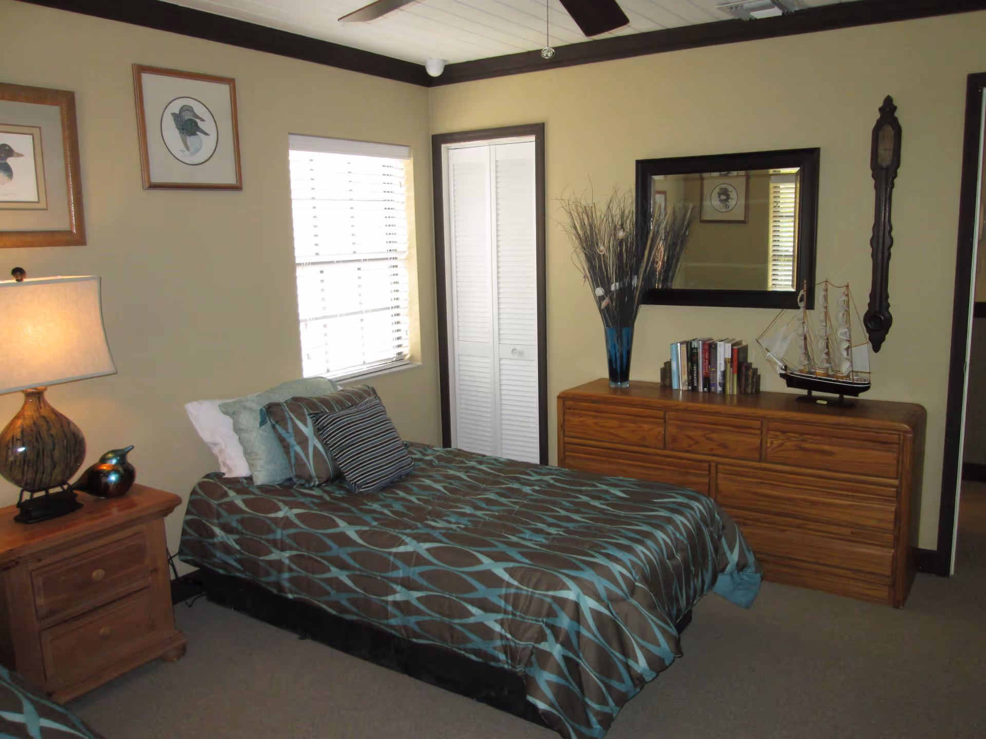 Bedroom with a single bed dressed in patterned teal and brown bedding, a nightstand with a lamp, and a wooden dresser topped with a mirror and decorative items.