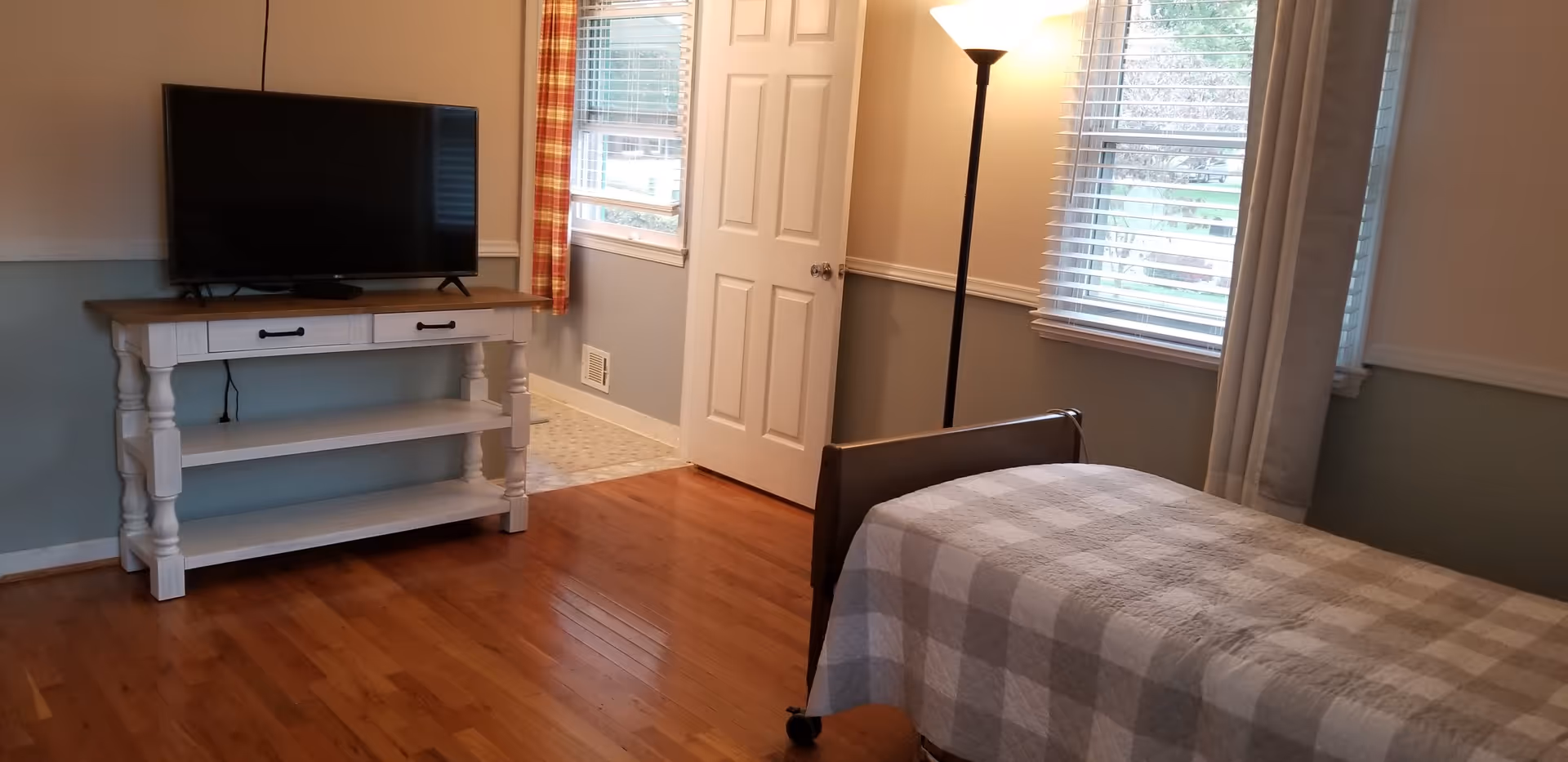 A bedroom with a single bed covered in a checkered blanket, a wooden floor, a white door slightly open, two windows with blinds and curtains, a floor lamp, and a TV on a white wooden stand.