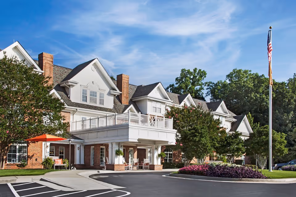 Exterior view of a senior living facility building with white siding and brick accents, featuring a covered entrance with white columns, rocking chairs, and a balcony above. There are trees, shrubs, and a flower bed with purple and pink flowers in front, along with a flagpole displaying the American flag. The sky is clear with some wispy clouds.