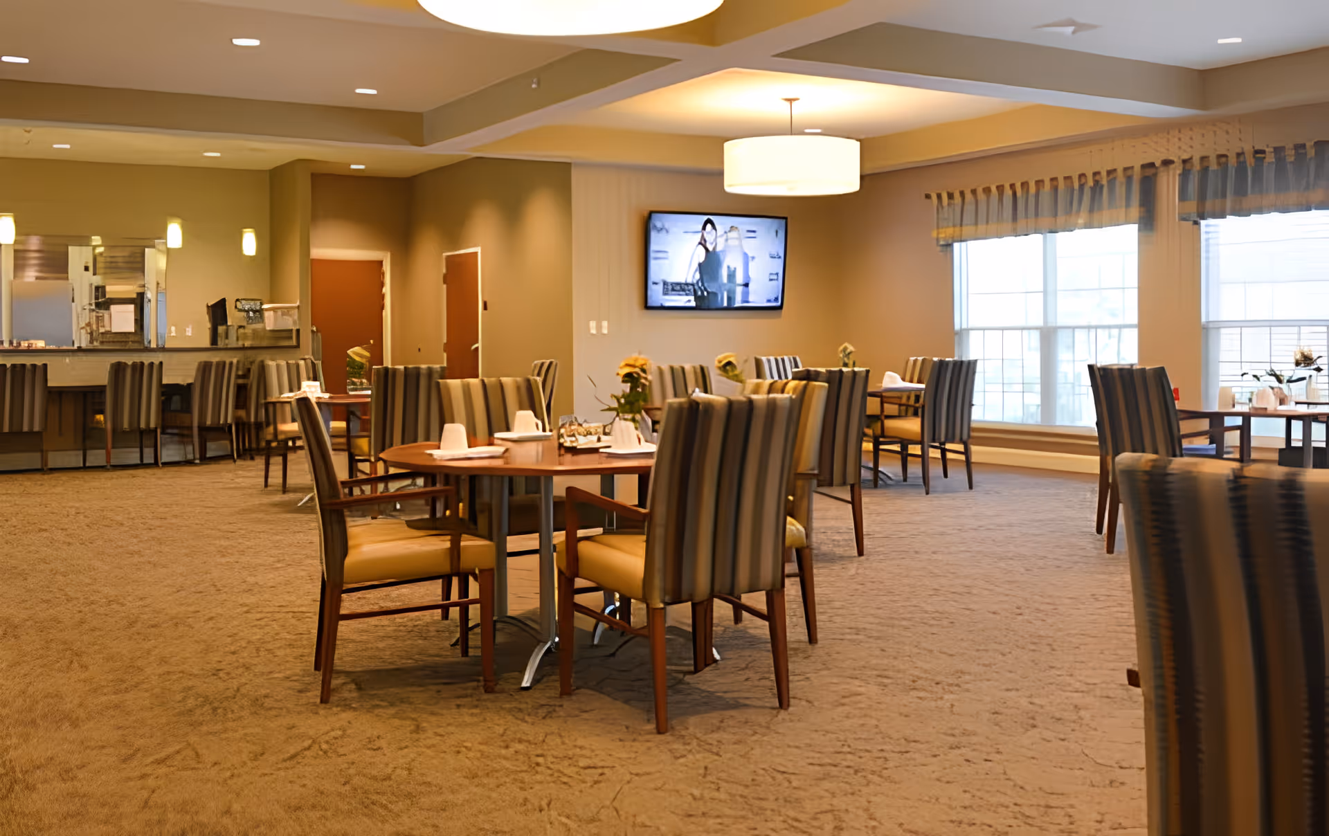Spacious dining room with round wooden tables and striped upholstered chairs, large windows, and a wall-mounted TV.