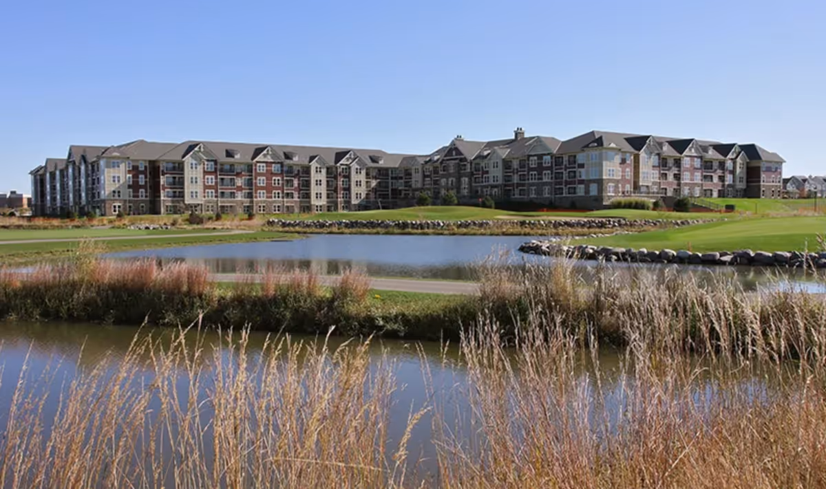 A large multi-story residential building with a mix of beige and red brick exterior, situated behind a pond and grassy area with tall dry grasses in the foreground under a clear blue sky.