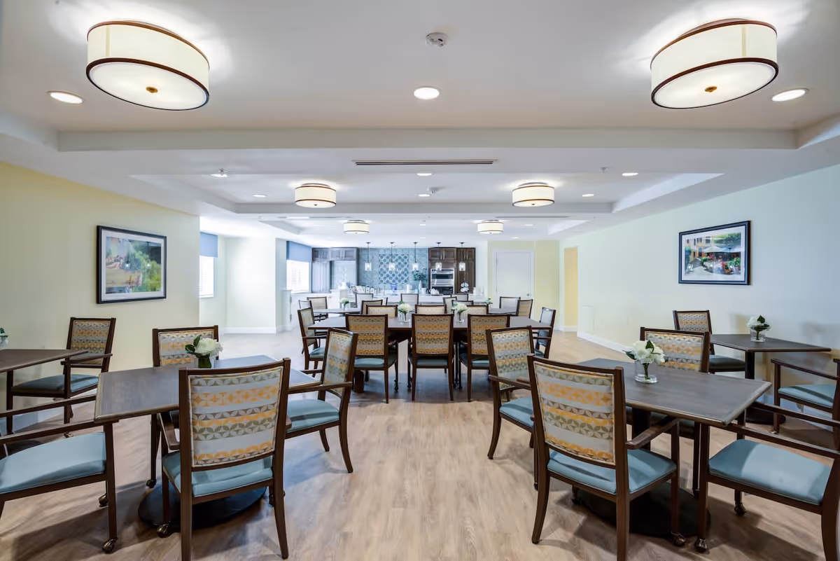 A bright and spacious dining room with multiple wooden tables and chairs arranged neatly. The chairs have patterned backs and light blue cushions. The room features light-colored walls, wood flooring, modern ceiling lights, and framed artwork on the walls. In the background, there is a kitchen area with dark cabinets and stainless steel appliances.