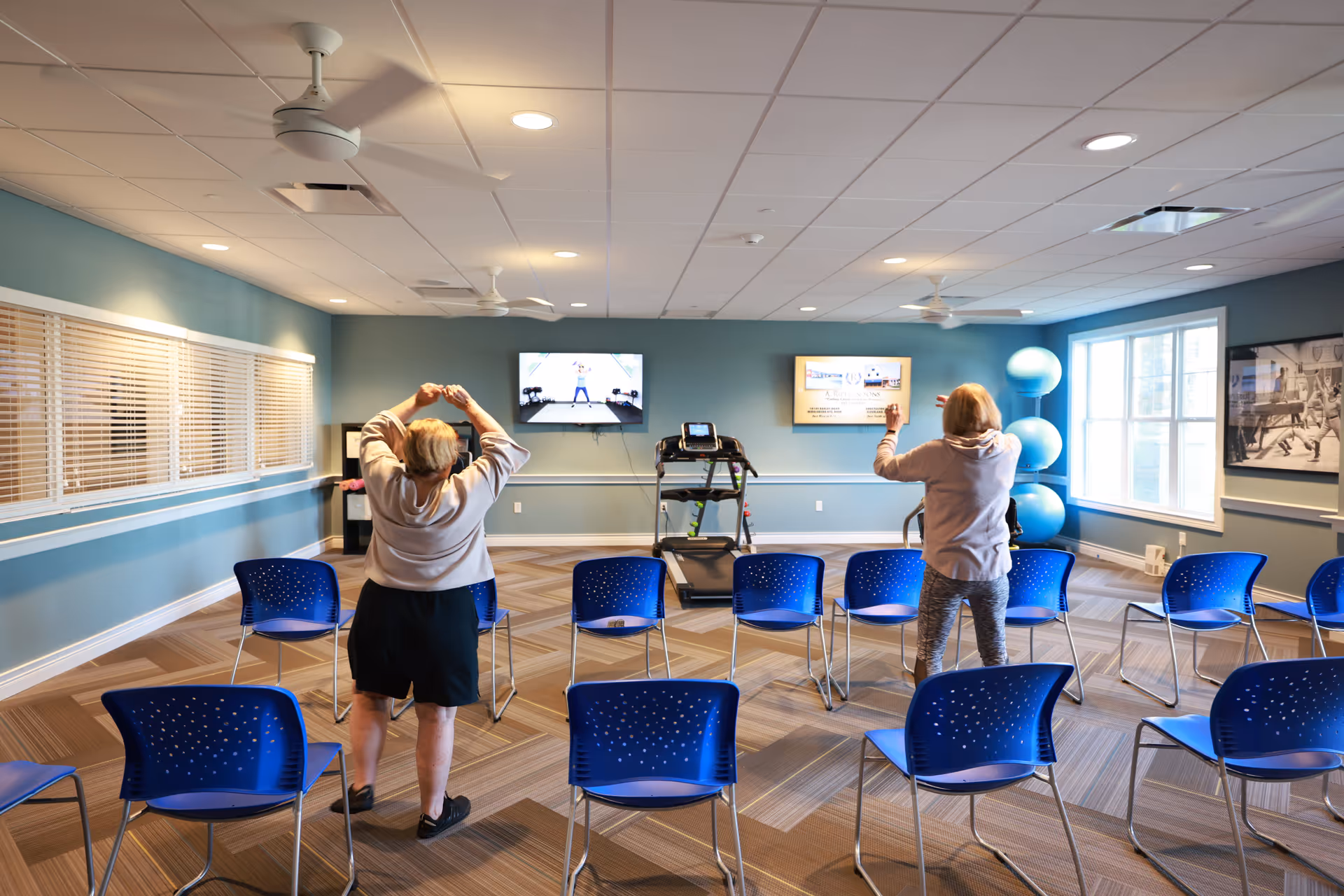 Two elderly women participating in a seated exercise class in a bright, spacious room with blue walls and blue chairs arranged in rows. They are following a workout video displayed on a wall-mounted TV. The room has large windows, ceiling fans, and exercise balls stacked in the corner.