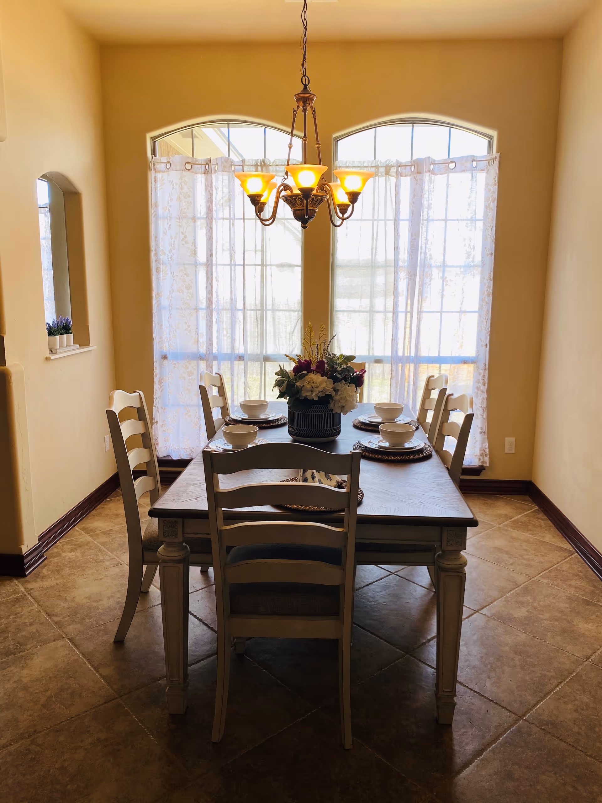 Dining room with a wooden table set for six, a floral centerpiece, and a chandelier in front of large arched windows with sheer curtains.