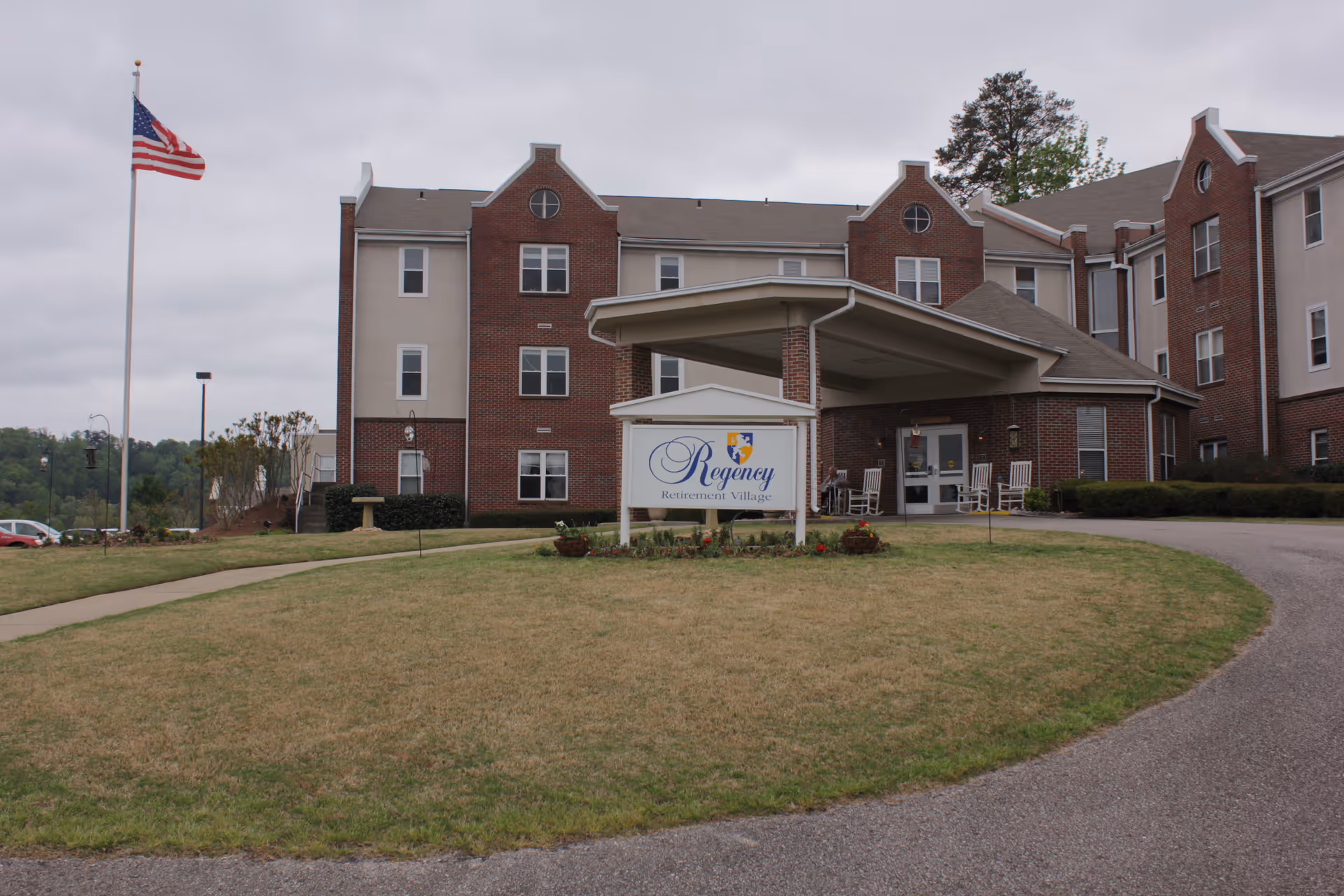 Exterior view of Regency Retirement Village of Birmingham, showing a multi-story brick and beige building with a covered entrance. There is a sign in front of the building with the facility's name, a curved driveway, a grassy lawn, and an American flag on a flagpole to the left.