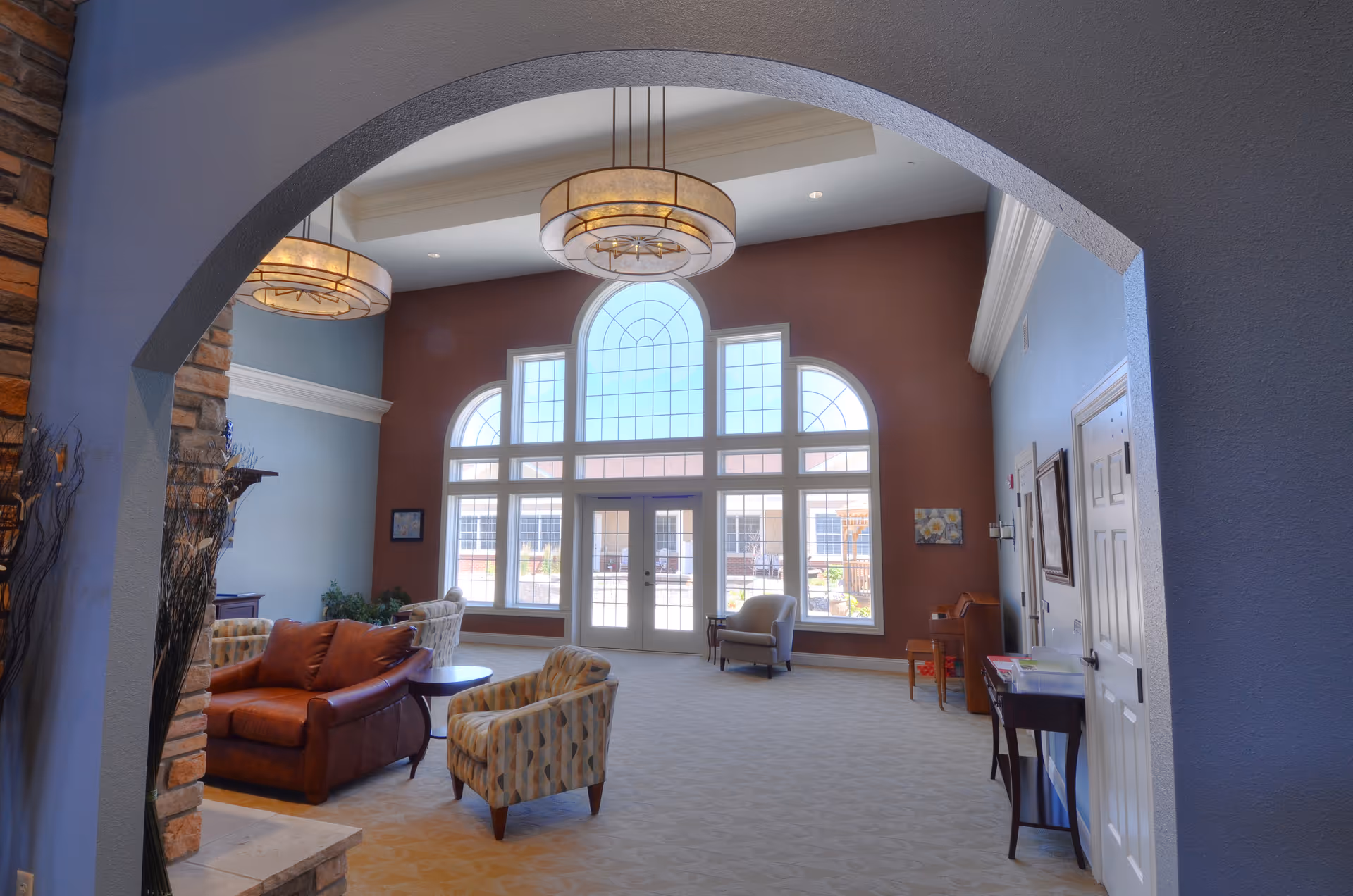 A spacious and well-lit living room area with large arched windows letting in natural light. The room features comfortable seating including a brown leather sofa and patterned armchairs, a small round table, and decorative hanging light fixtures. The walls are painted in warm tones with a mix of blue and brown, and there is a stone fireplace partially visible on the left side.