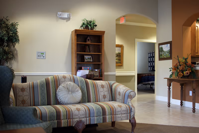 A cozy living room area with a striped upholstered sofa and a round decorative pillow. To the left is a green armchair partially visible. Behind the sofa is a wooden bookshelf with some books and decorative items. The room has beige walls with white trim and a potted plant in the corner. An arched doorway leads to a hallway and another room with a bed visible. A wooden table with a floral arrangement is on the right side near another archway leading to a kitchen area.