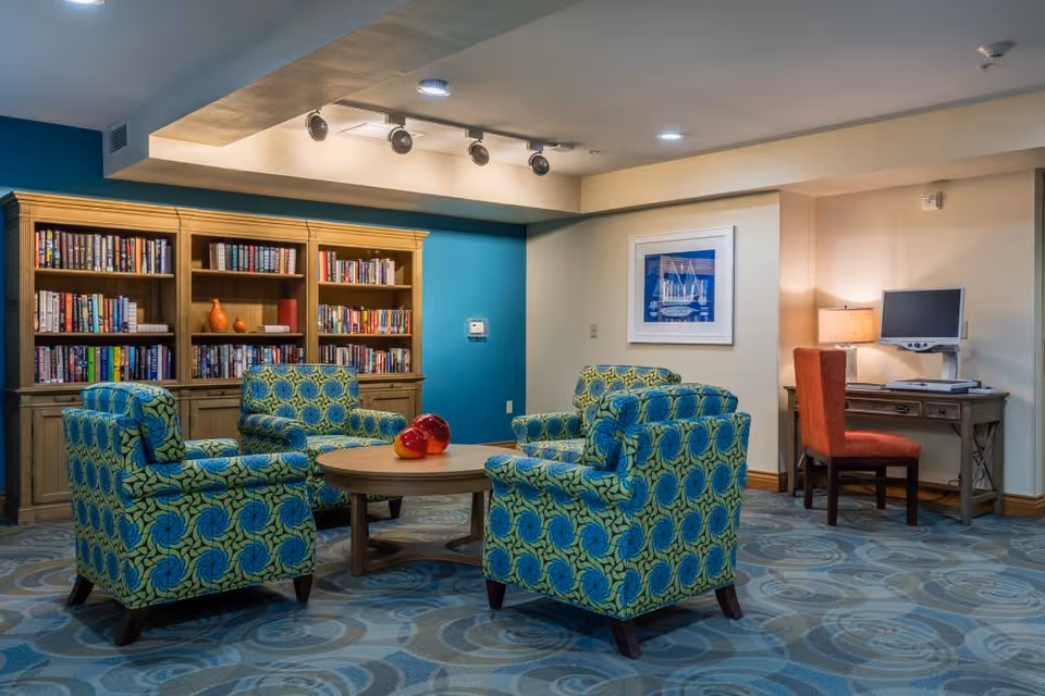 Cozy seating area with four patterned armchairs around a round wooden coffee table, a bookshelf wall, and a small desk with a computer.