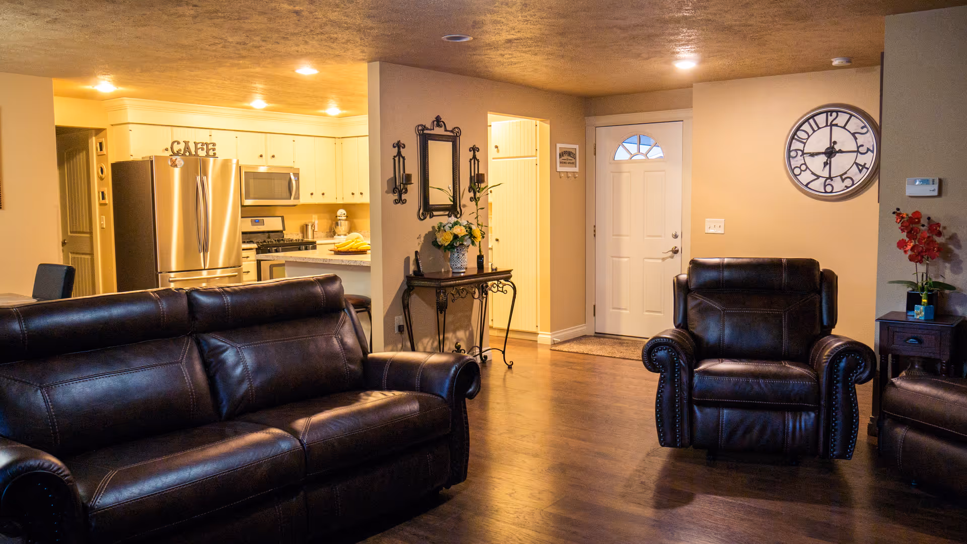 Interior view of a living room and kitchen area in a senior living facility. The living room features dark brown leather sofa and armchairs on a wooden floor. The kitchen has white cabinets, a stainless steel refrigerator, microwave, and stove. A small table with a flower vase and a mirror is placed near the entrance door. A large wall clock is mounted on the wall above a side table with a red flower arrangement.