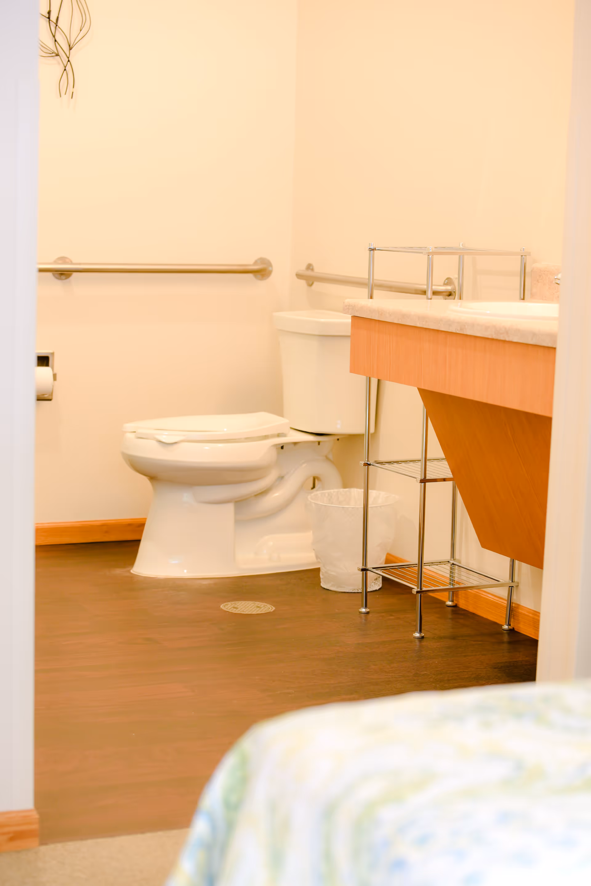 View of a clean bathroom with a white toilet, grab bars on the walls, a wooden vanity with a countertop, a small metal shelving unit, and a wastebasket. The floor is dark wood, and part of a bed with a patterned bedspread is visible in the foreground.