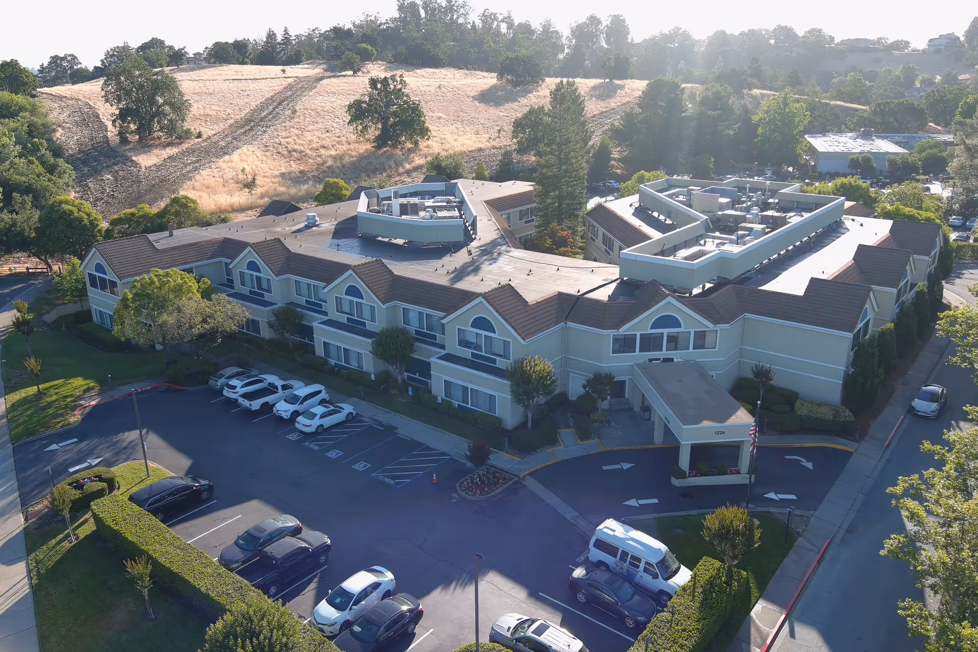 Aerial view of a two-story senior living/post-acute building with a covered entrance, parking lot, and surrounding trees and hills.