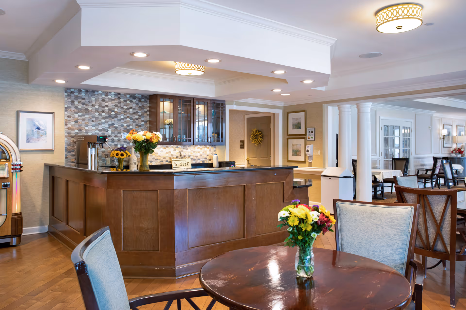 Well-lit bistro-style dining area with a wooden service counter, tables and chairs, and vases of fresh flowers.