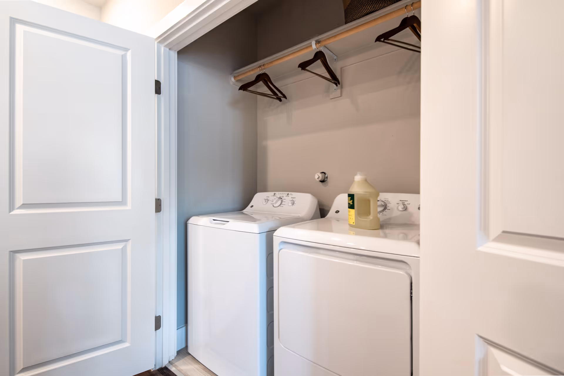 Laundry area with a white washing machine and dryer side by side inside a small closet space. There are a few empty hangers on a rod above the machines and a bottle of detergent on top of the dryer. The closet doors are open, revealing the appliances.