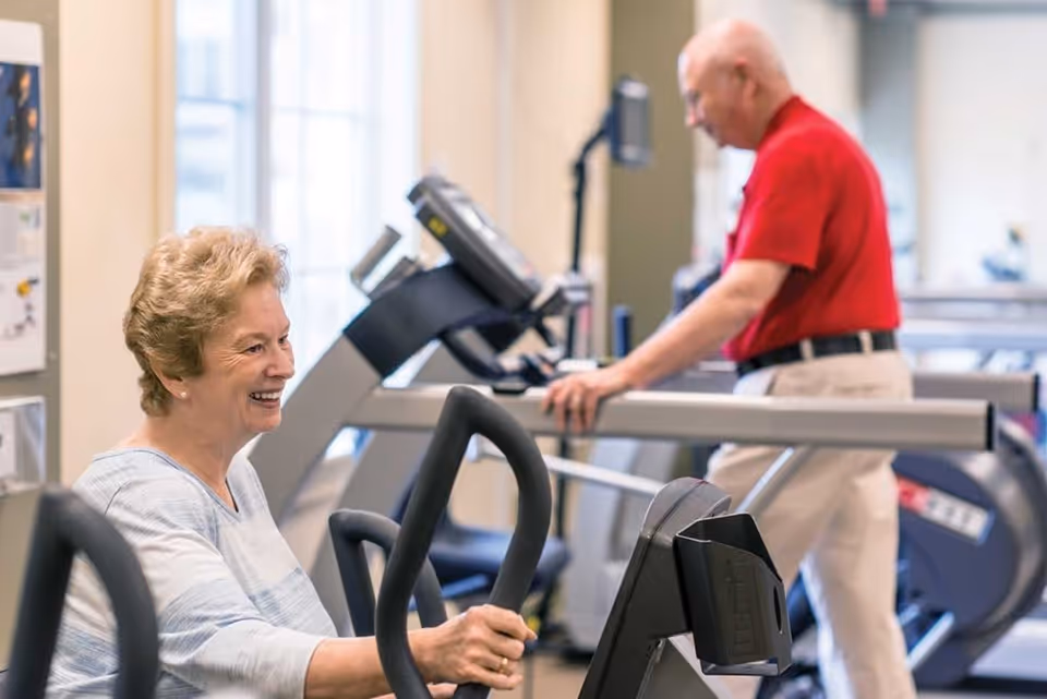 An elderly woman smiling while using a stationary exercise bike in a fitness room. In the background, an elderly man in a red shirt is walking on a treadmill. The room is well-lit with large windows.