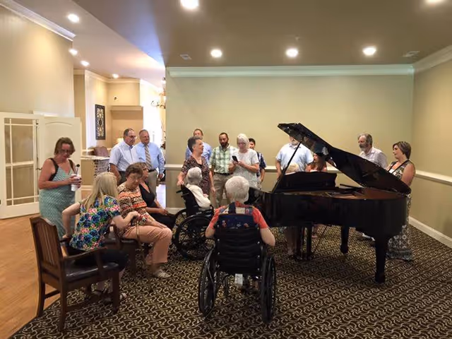 A group of seniors and staff gathered around a grand piano in a common room, with some people seated in chairs and wheelchairs and others standing.