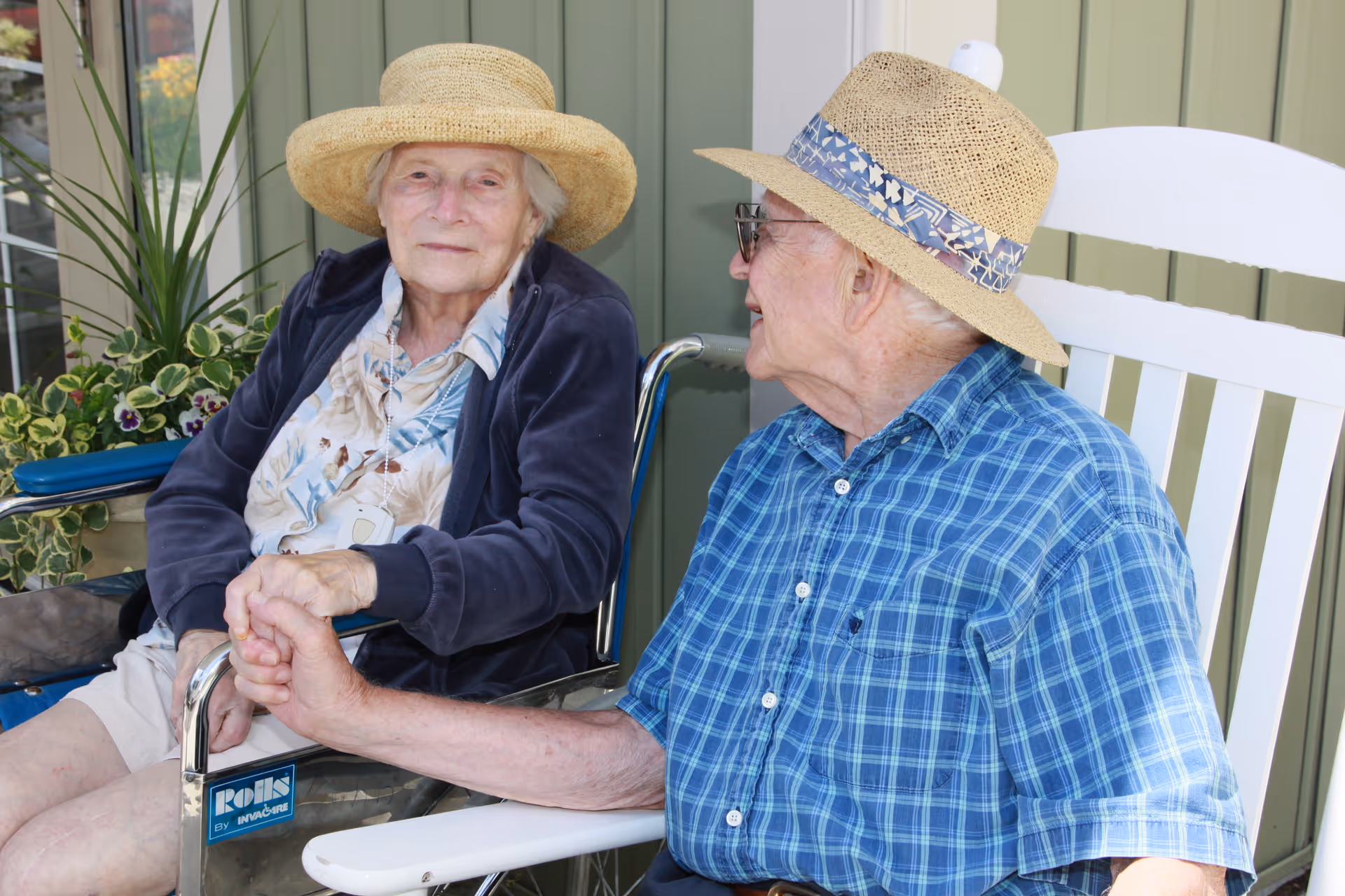 Two elderly individuals wearing straw hats sitting outdoors. One person is in a wheelchair holding hands with the other person who is seated in a white rocking chair. There are green plants and a light green wall in the background.