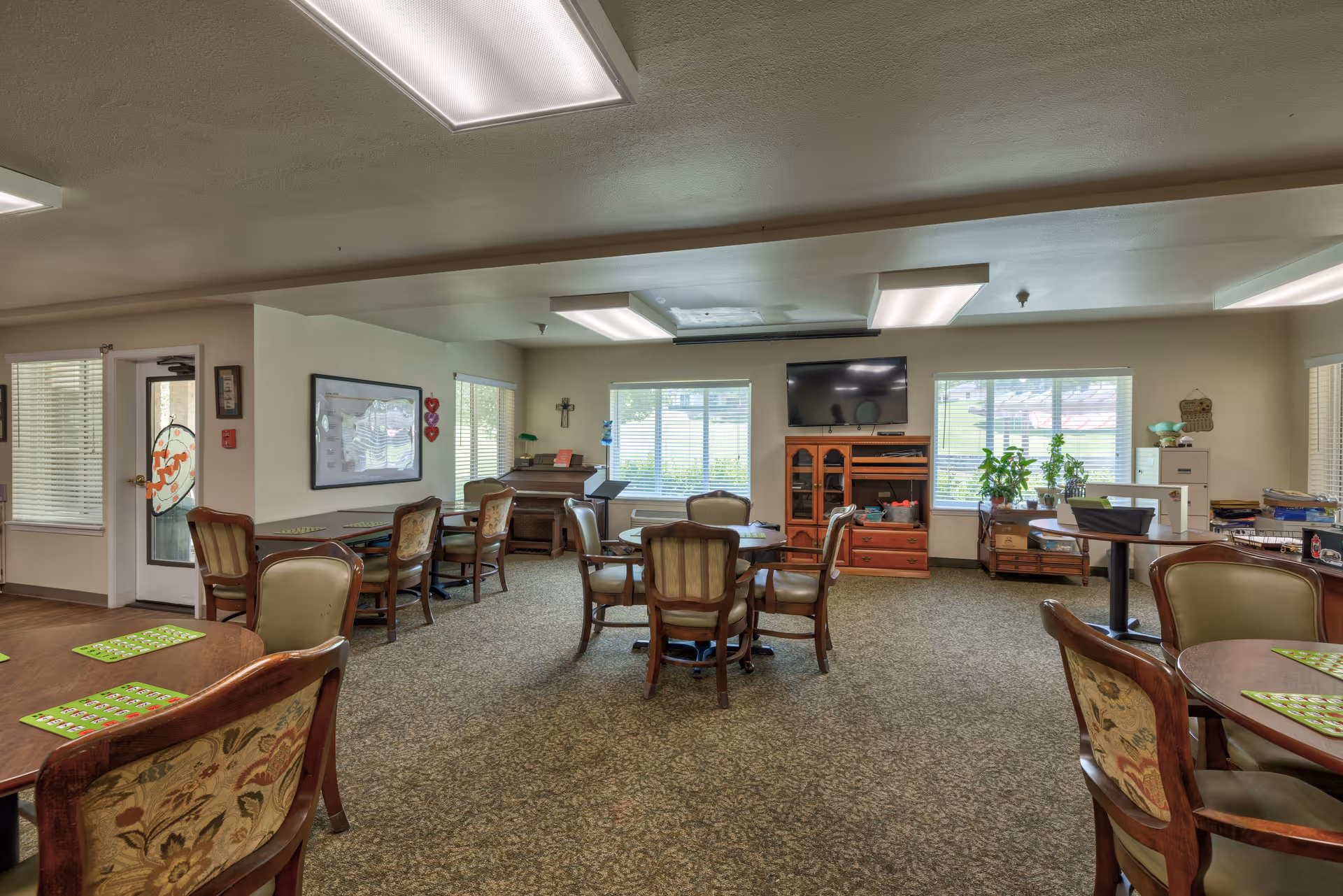 A well-lit common area in a senior living facility with multiple round tables and cushioned chairs arranged for social activities. The room features large windows with blinds, a television mounted on the wall above a wooden cabinet, a piano, and various plants and decorations. The carpeted floor and neutral wall colors create a warm and inviting atmosphere.