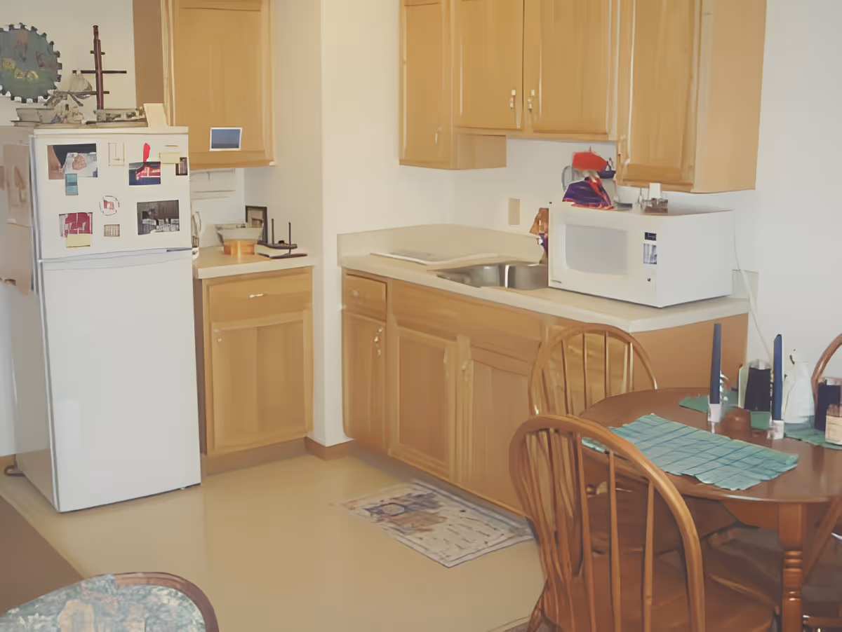 A small kitchen area with wooden cabinets, a white refrigerator with magnets and photos, a white microwave on the countertop, a stainless steel sink, and a wooden dining table with chairs. The table has a green placemat and two blue candles.