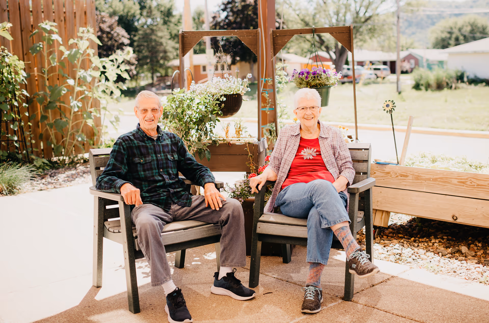 An elderly man and woman sitting outdoors on wooden chairs in a garden area with hanging flower pots and greenery around them, smiling at the camera on a sunny day.