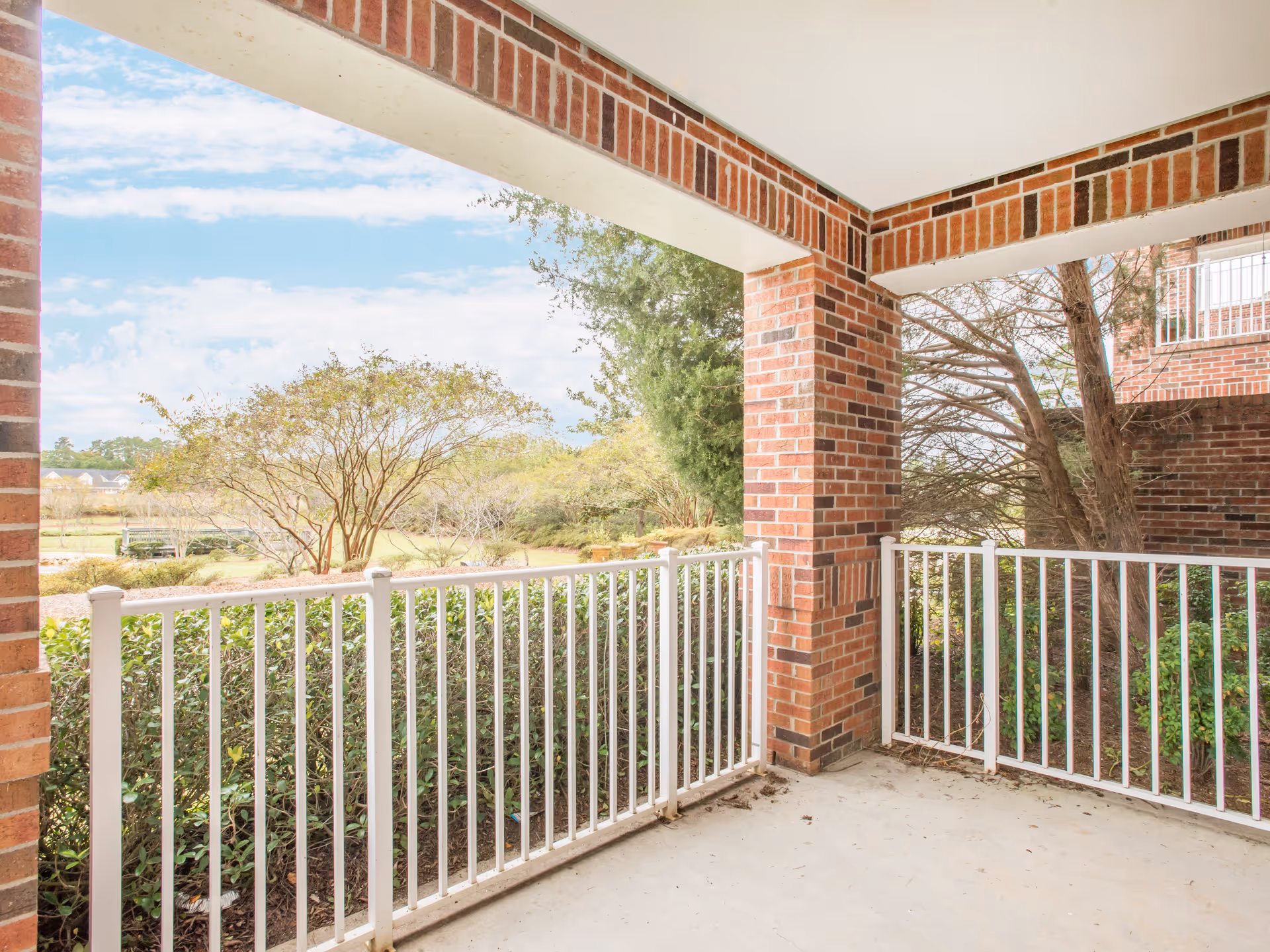 Covered outdoor patio area with white metal railing and brick pillars, overlooking greenery and trees under a partly cloudy sky.
