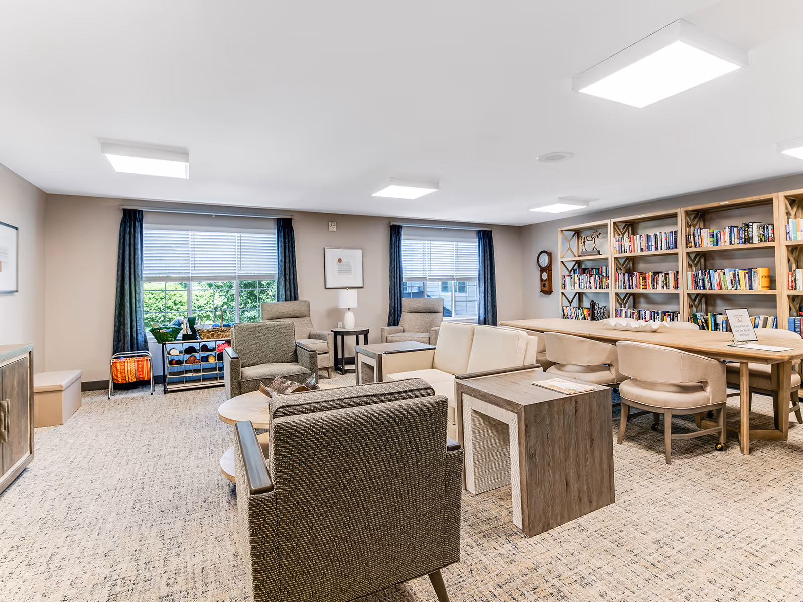 A bright and spacious common area in a senior living facility with comfortable seating including armchairs and a sofa, a large wooden table with chairs, bookshelves filled with books, two large windows with blue curtains, and a carpeted floor.
