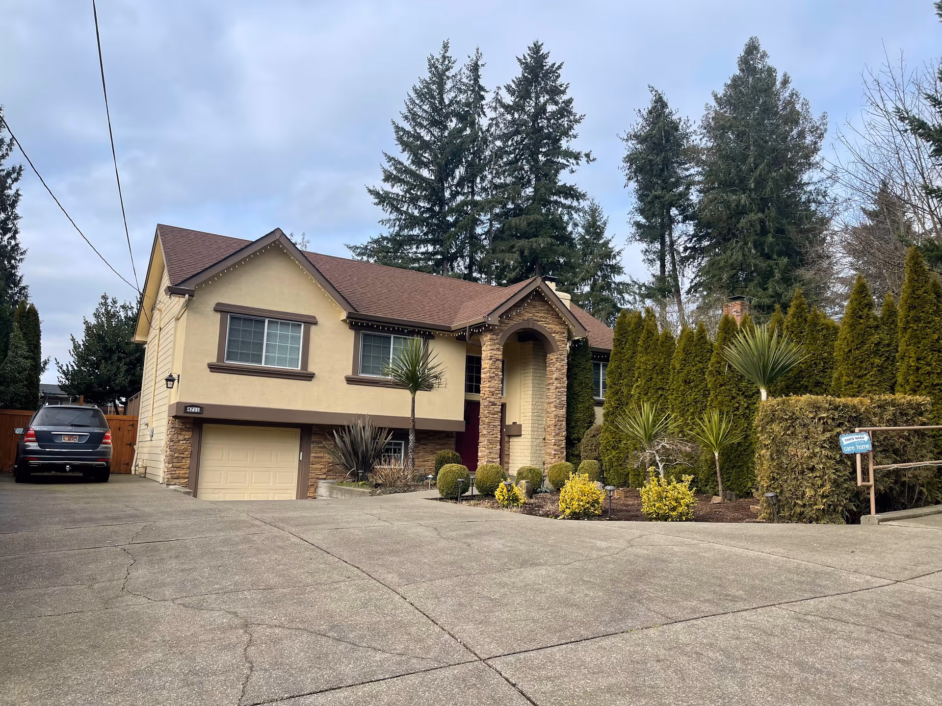 Exterior view of a two-story residential-style building with beige walls, brown roof, and stone accents around the entrance. There is a driveway leading to a garage, a black SUV parked on the left side, and well-maintained landscaping with bushes and tall trees surrounding the property. A small sign near the right side of the driveway indicates it is a care home.