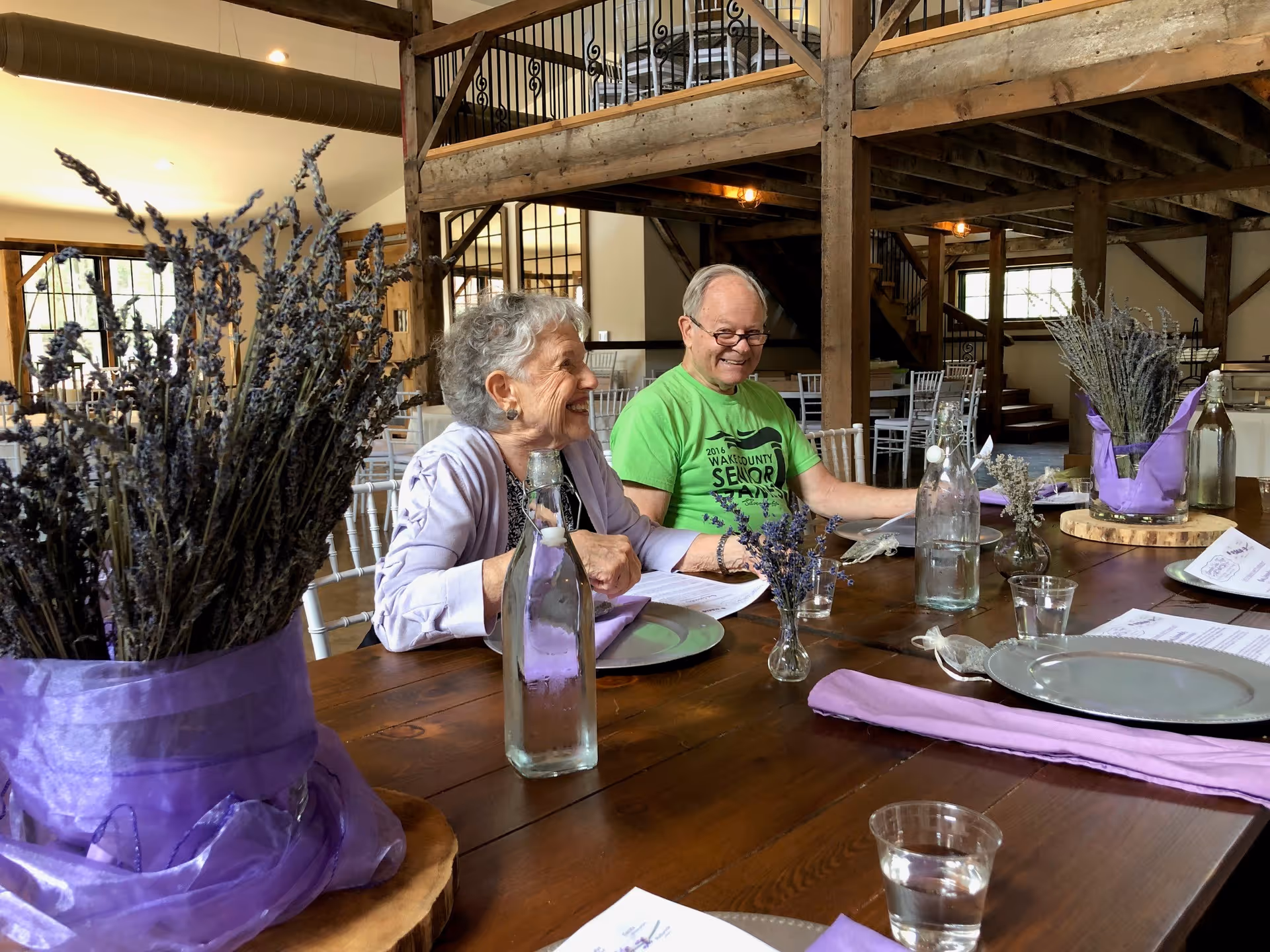 Two elderly people sitting at a wooden dining table in a rustic room with exposed wooden beams. The table is set with plates, glasses, water bottles, and lavender flower arrangements wrapped in purple fabric. The people are smiling and appear to be enjoying a conversation.