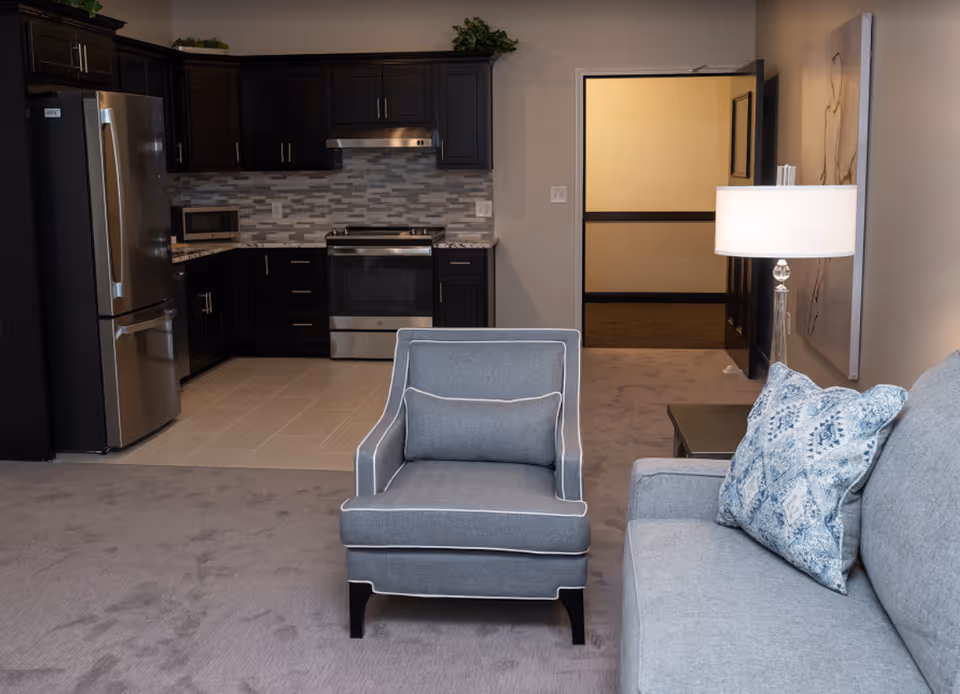 Interior view of a senior living facility showing a small kitchen area with dark wood cabinets, stainless steel refrigerator, stove, and microwave. In the foreground, there is a gray armchair with a matching pillow and part of a gray sofa with a blue patterned pillow. A table lamp is on a side table next to the sofa, and an open doorway leads to a hallway.