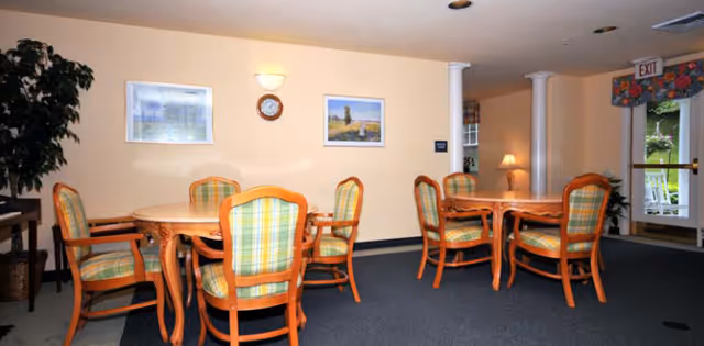 Interior view of a common area with two wooden tables surrounded by eight wooden chairs with green and yellow plaid cushions. The room has beige walls with two framed pictures and a clock, a potted plant in the corner, a lamp on a side table, and an exit door with a floral valance above it.
