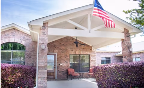 Front exterior view of a brick building with a covered entrance supported by brick columns. There is an American flag mounted on the roof above the entrance, two chairs and a small table under the covered area, and bushes on either side of the walkway leading to the door.