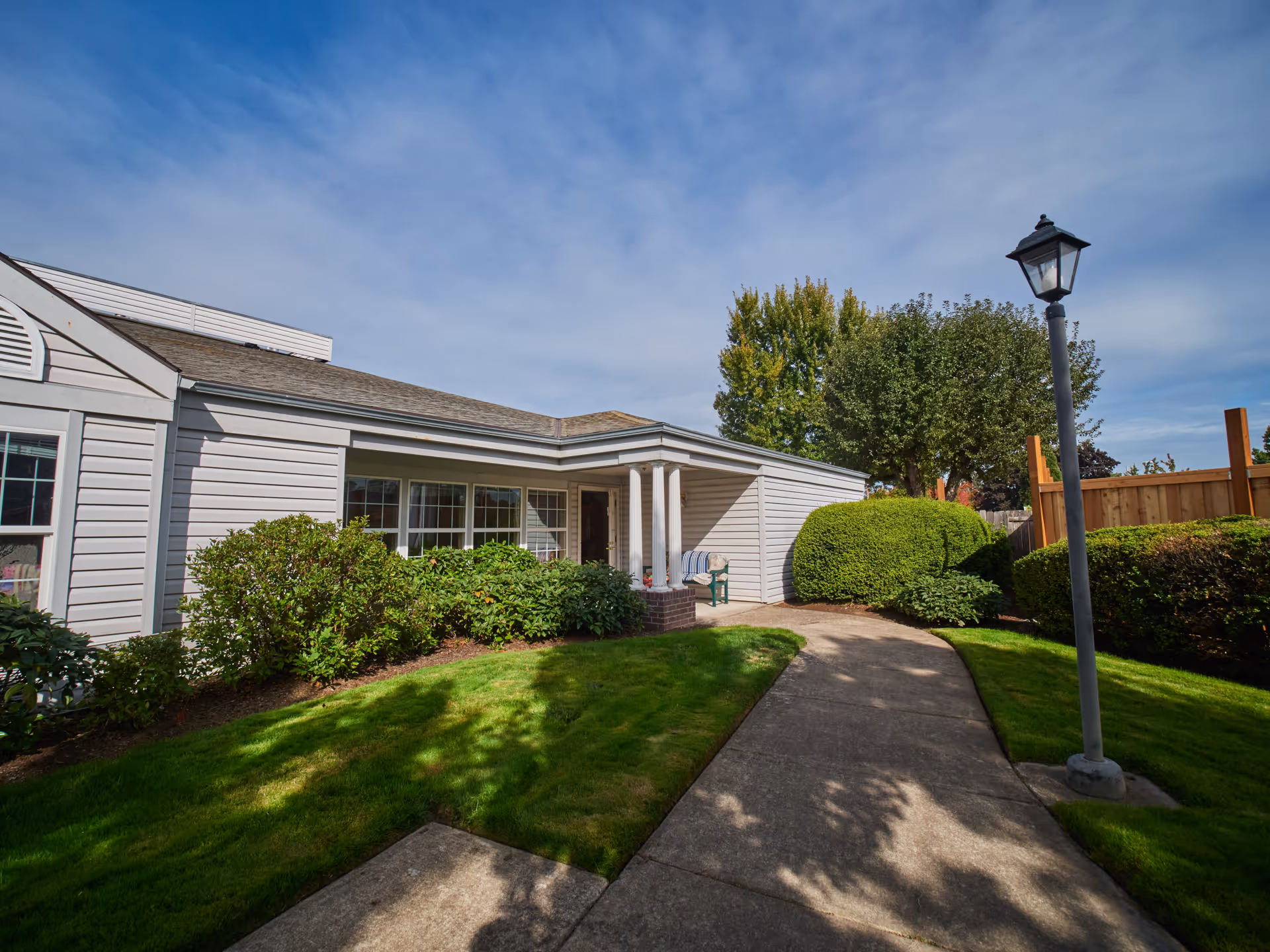 Exterior view of a single-story senior living facility building with light gray siding, large windows, and a covered entrance supported by white columns. A concrete pathway leads to the entrance, surrounded by well-maintained green grass, bushes, and a lamppost. The sky is clear with some clouds.