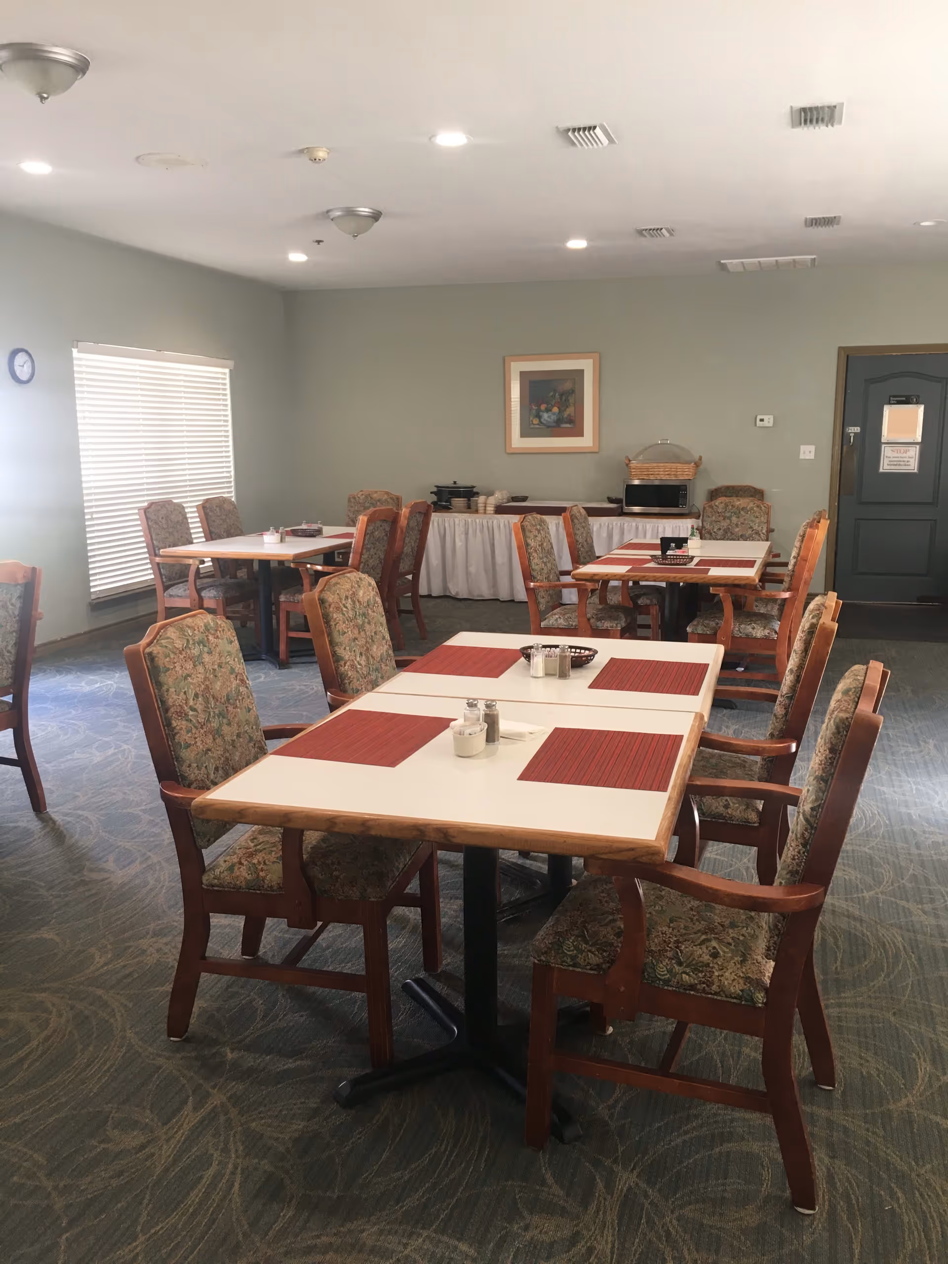 A dining room with several tables and chairs arranged neatly. Each table has red placemats and condiments in the center. The room has carpeted flooring, light green walls, a window with blinds, and a door at the back. A framed picture hangs on the wall above a buffet table covered with a white tablecloth holding some kitchen appliances and baskets.