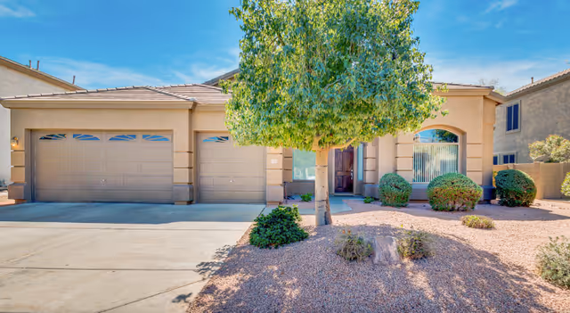 Front exterior view of a single-story house with a three-car garage, a tree, and desert landscaping with gravel and bushes under a blue sky.