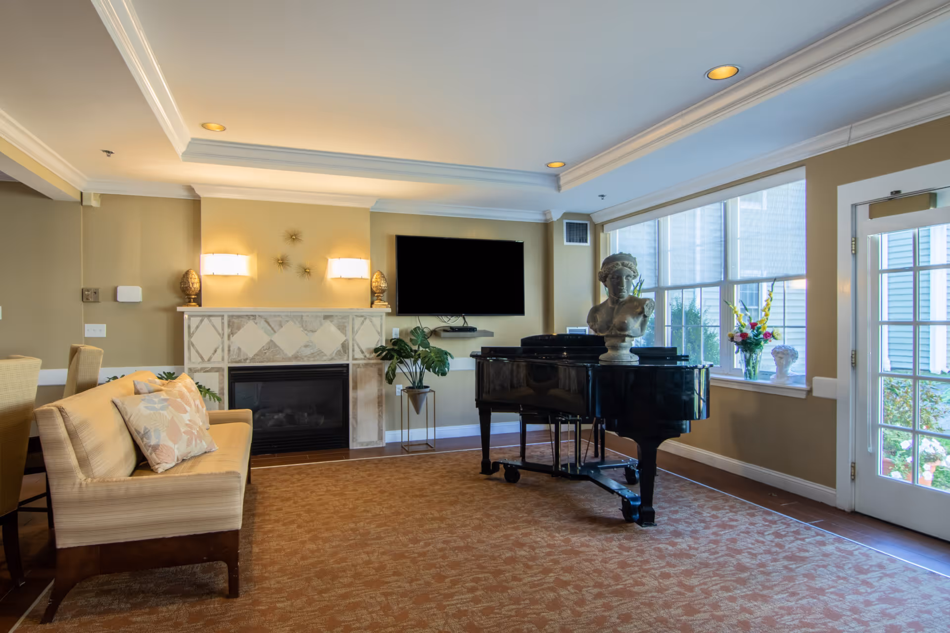 Sunny communal living room with a grand piano, fireplace, wall-mounted TV, seating, and large windows.