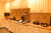 A kitchen area with white cabinets and drawers, a granite countertop, a coffee maker, a kettle, a bowl of fruit, and a tiled backsplash.