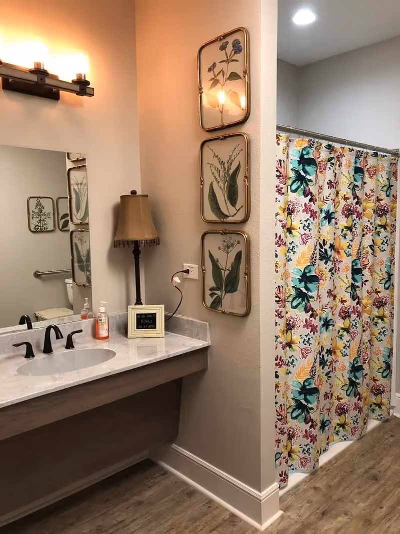 A bathroom interior featuring a marble countertop with a sink and black faucet. On the countertop, there is a soap dispenser, a small framed sign, and a decorative lamp. Above the sink is a large mirror with a light fixture above it. On the wall next to the countertop, there are three framed botanical prints arranged vertically. To the right, there is a shower area with a colorful floral shower curtain. The floor has a wood-like finish.