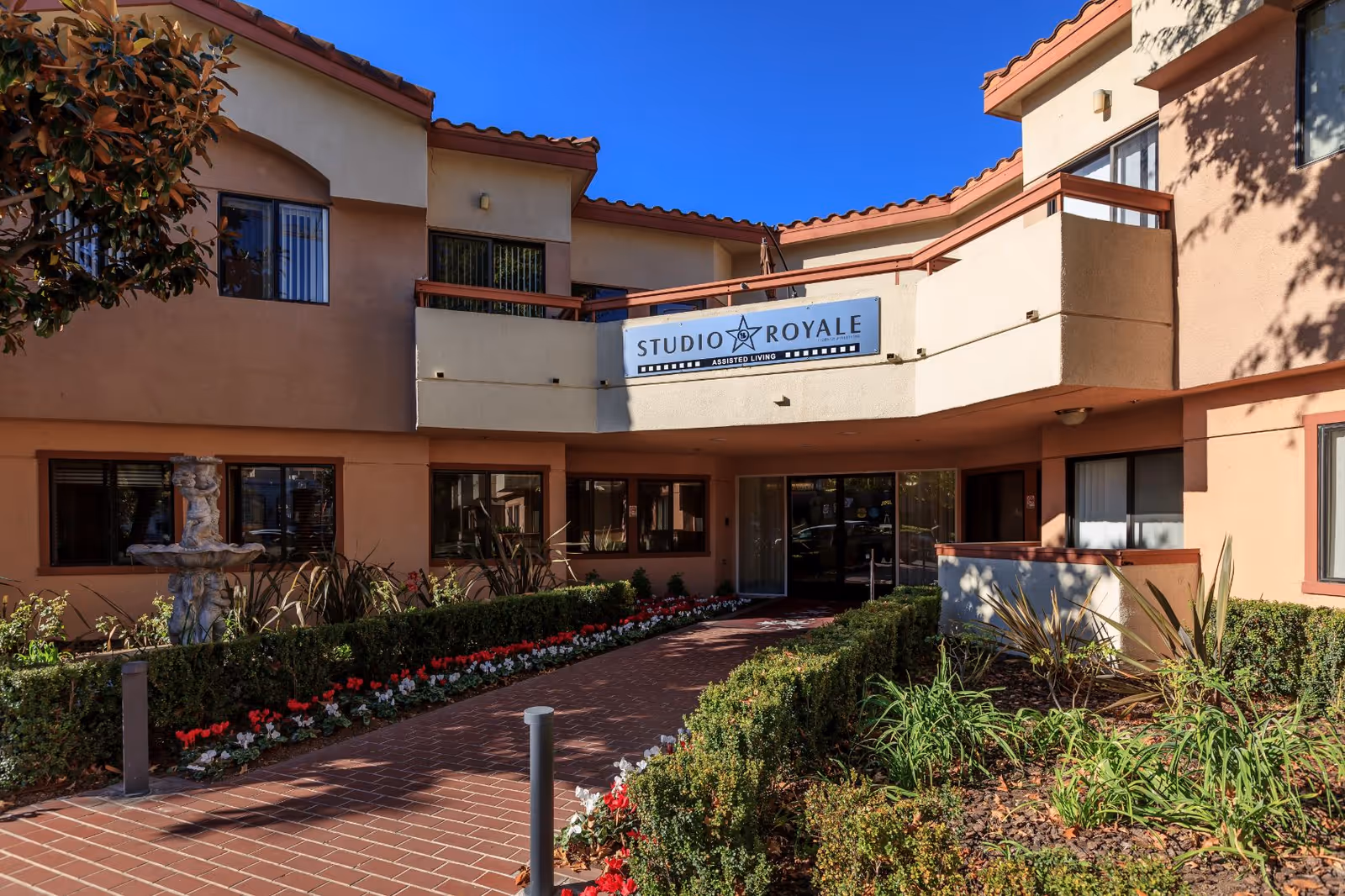 Exterior view of Studio Royale assisted living facility with a walkway lined by bushes and flowers leading to the entrance. The building has beige walls with red trim and a sign above the entrance that reads 'Studio Royale Assisted Living'.