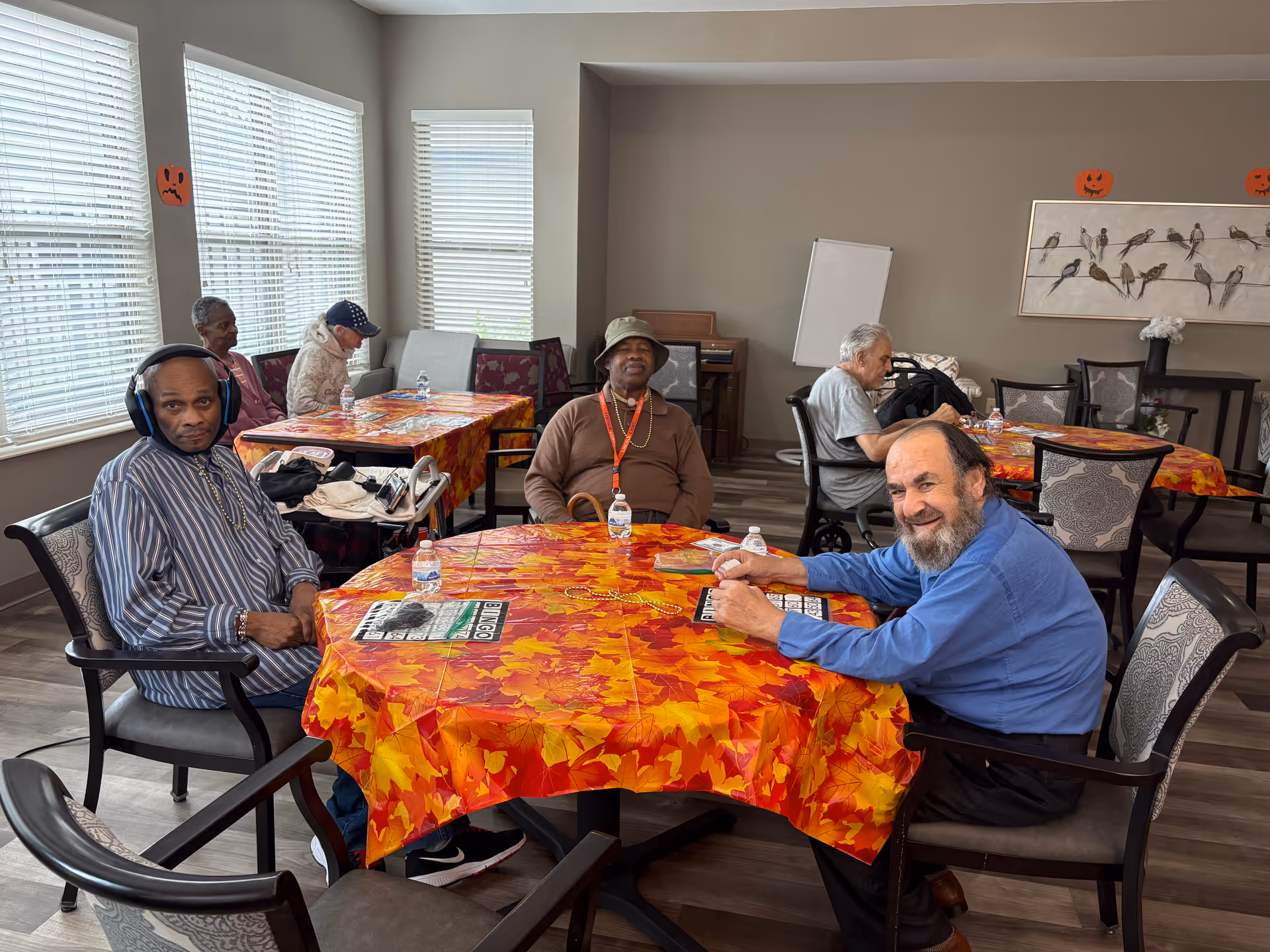 Several older adults seated around tables with autumn leaf tablecloths in a communal activity room.