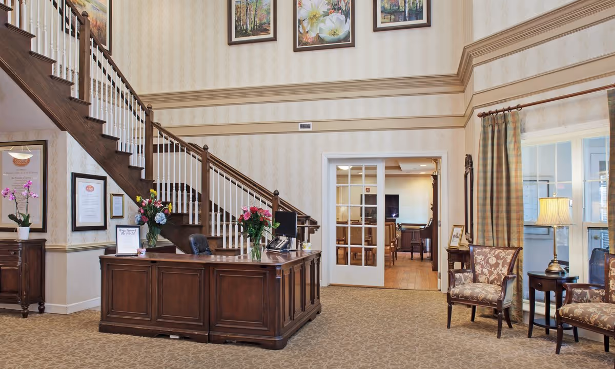 Sunlit lobby with a wooden reception desk, grand staircase, floral arrangements and a seating area with chairs and lamps.