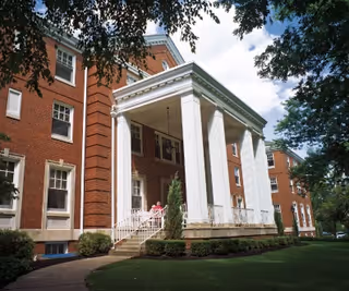 Exterior view of a large brick building with white columns at the entrance, surrounded by green grass and trees under a partly cloudy sky.