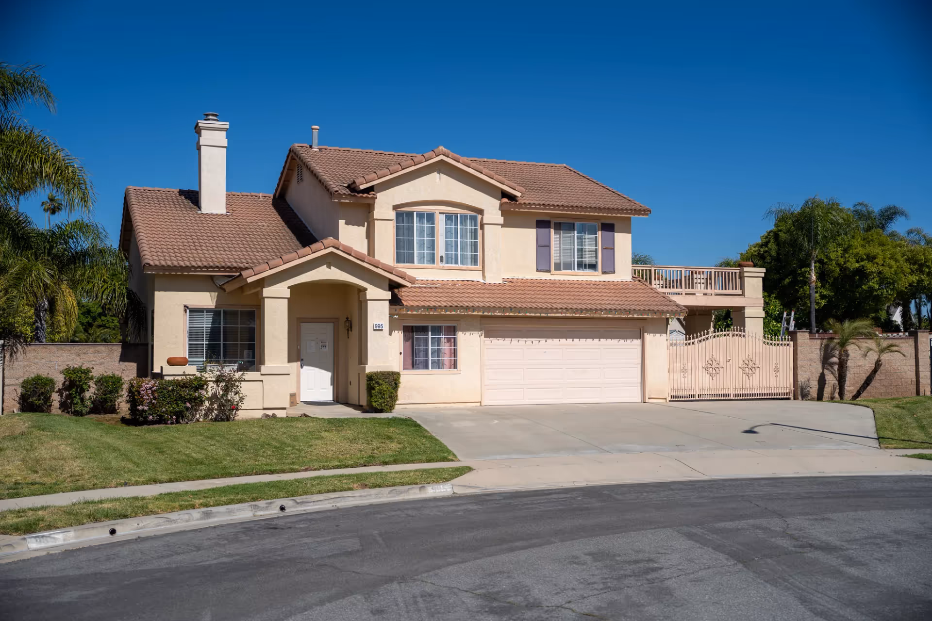 Two-story beige suburban house with tiled roof, driveway, and gated front yard.