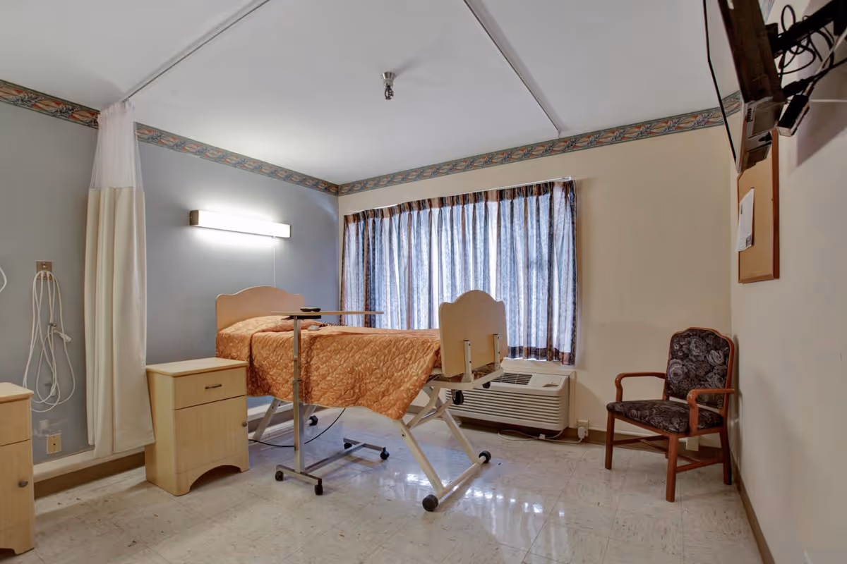 Small clinical patient bedroom with an adjustable hospital bed, over-bed table, nightstand, chair, and a curtained window.