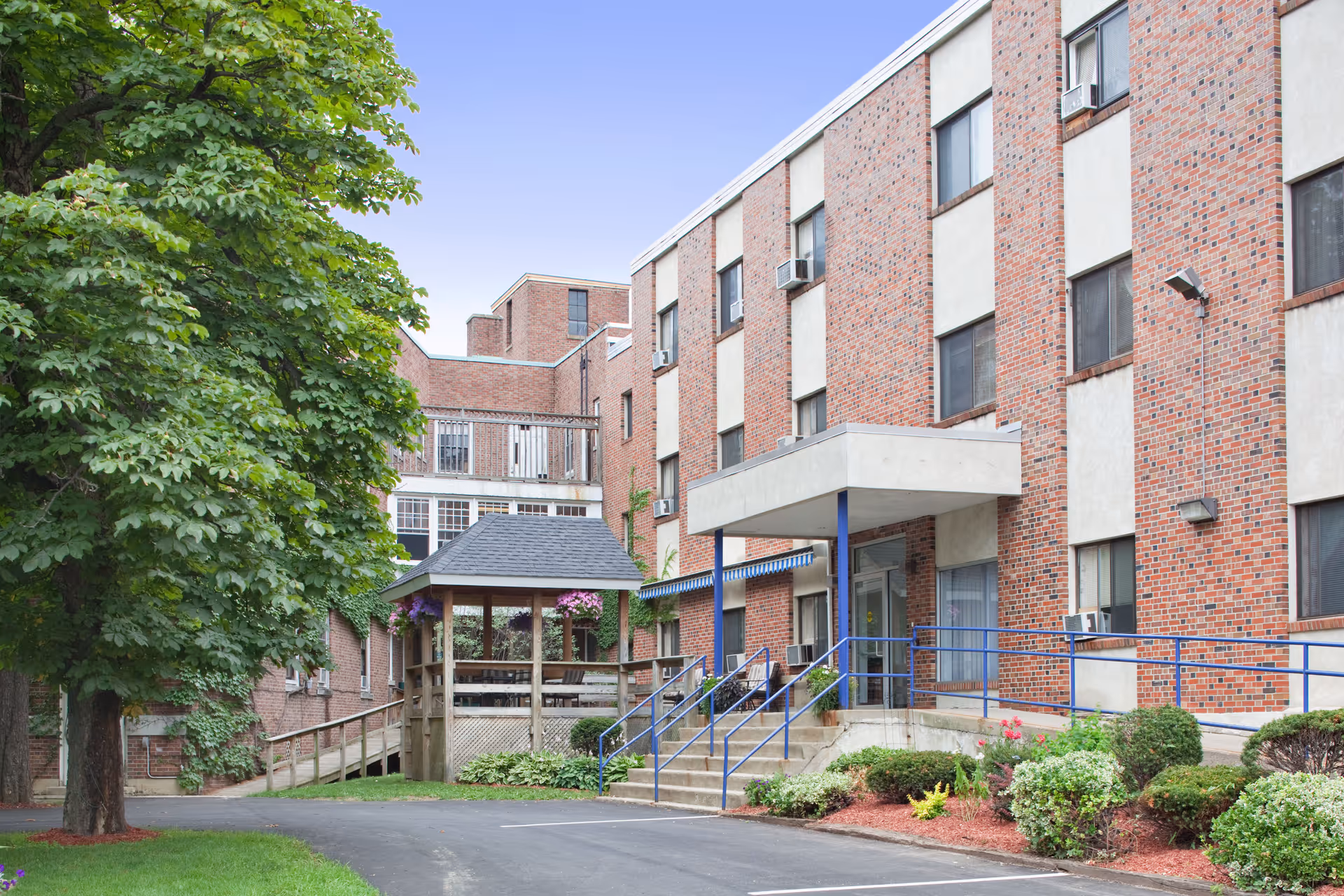 Brick multi-story senior living facility front with an entrance canopy, blue handrails, a gazebo, ramp, and landscaped grounds.