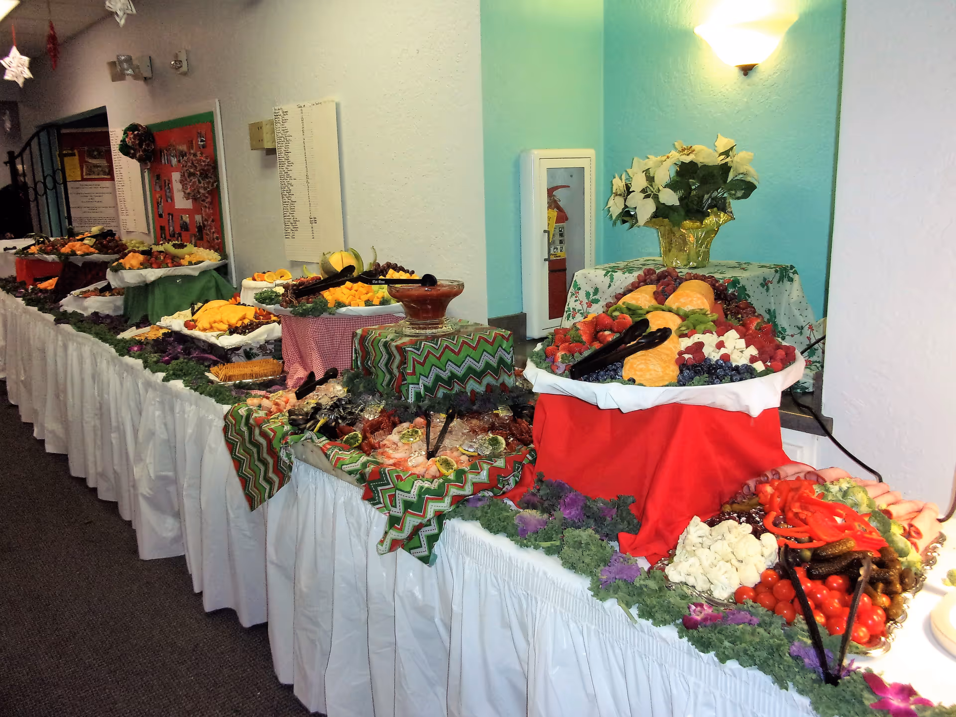 A long buffet table covered with white tablecloth and decorated with green and red patterned cloths, displaying a variety of foods including fruits, vegetables, cheeses, crackers, and dips. The table is set against a wall with a light blue accent and a wall-mounted light fixture above a vase of white flowers. The setting appears to be indoors, possibly in a community or dining area.