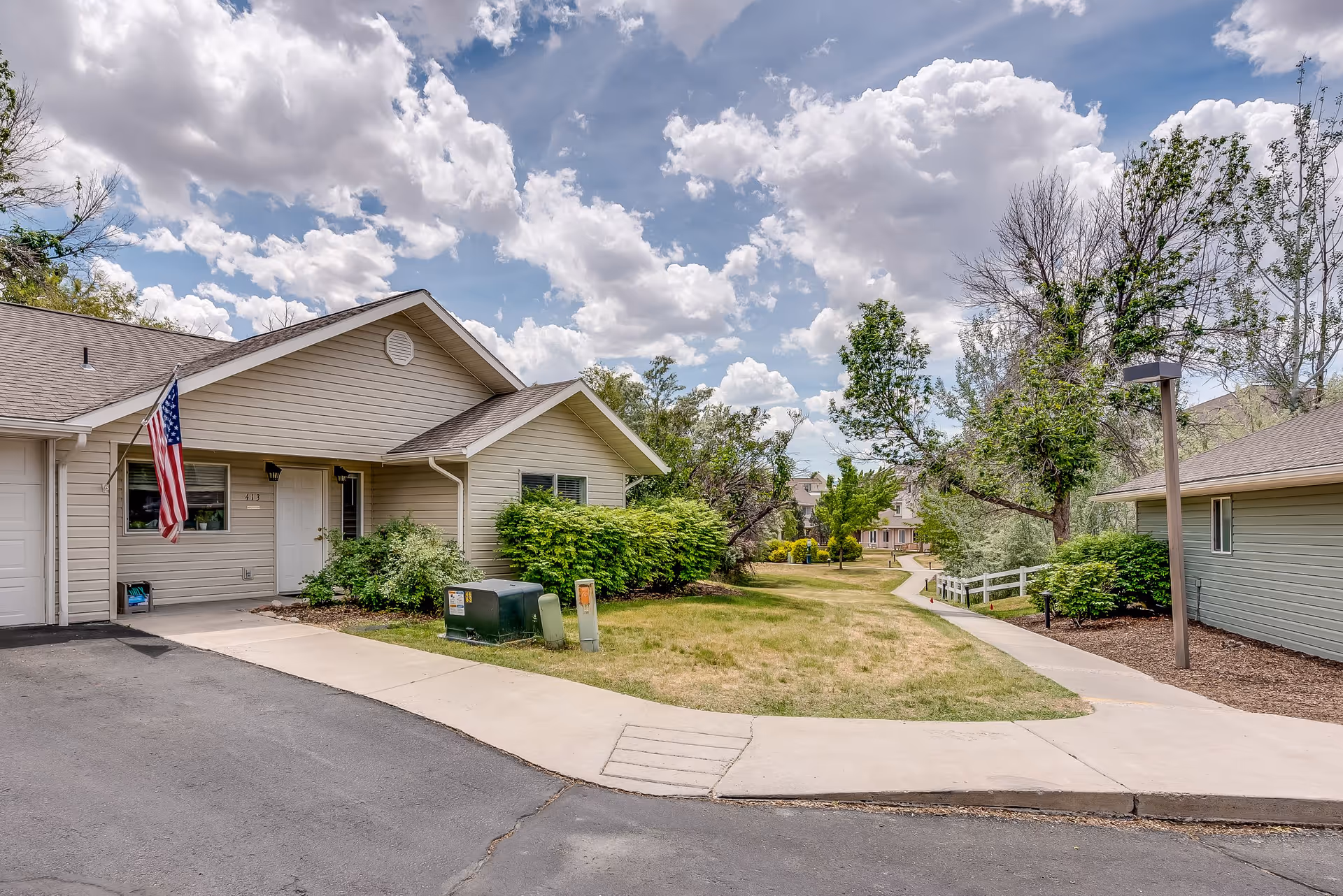 Exterior view of a senior living facility with beige siding buildings, a paved driveway, green bushes, trees, and a partly cloudy sky. An American flag is mounted near the entrance of one building.