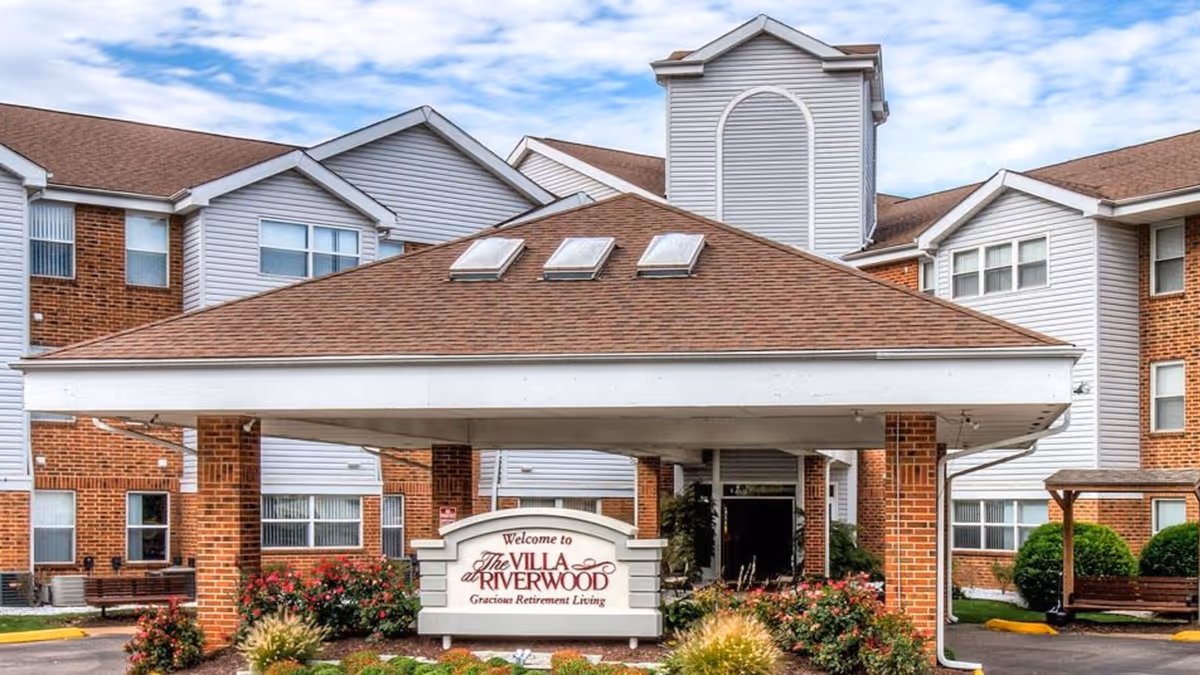 Exterior view of The Villa at Riverwood, a multi-story brick and siding building with a covered entrance supported by brick columns. There are landscaped bushes and flowers around the entrance, and a sign that reads 'Welcome to The Villa at Riverwood Gracious Retirement Living'.
