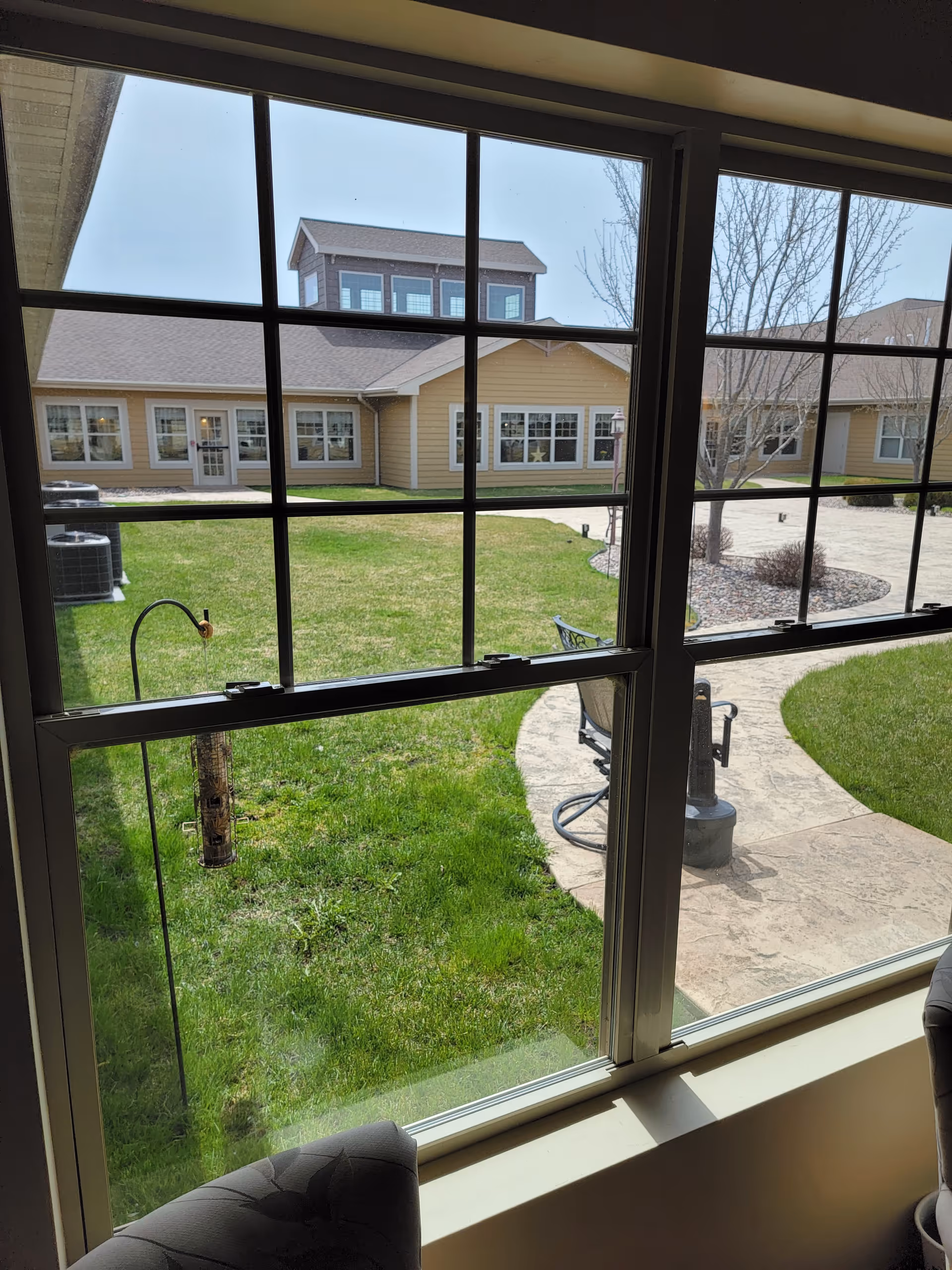 View through a large window showing a green lawn, a curved concrete pathway, outdoor chairs, a bird feeder, and a beige building with multiple windows under a clear sky.