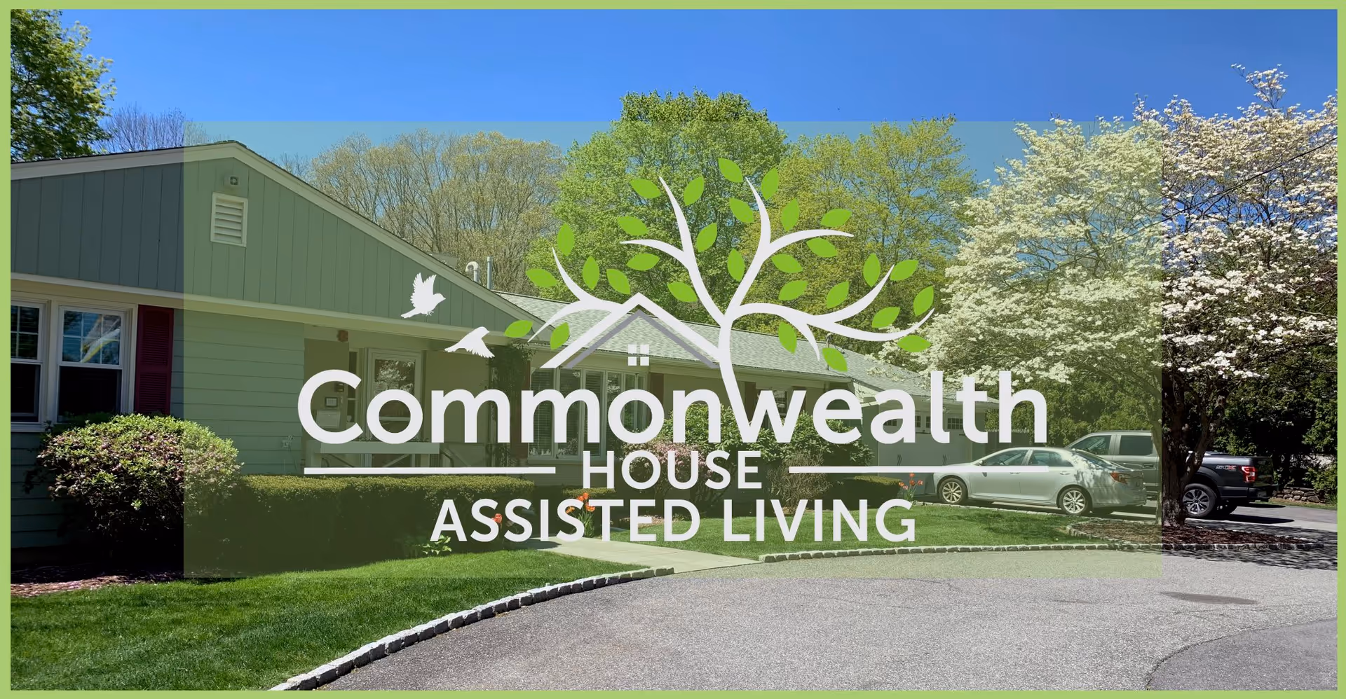 Exterior view of a single-story assisted living facility building surrounded by green trees and bushes, with a driveway and parked cars visible.