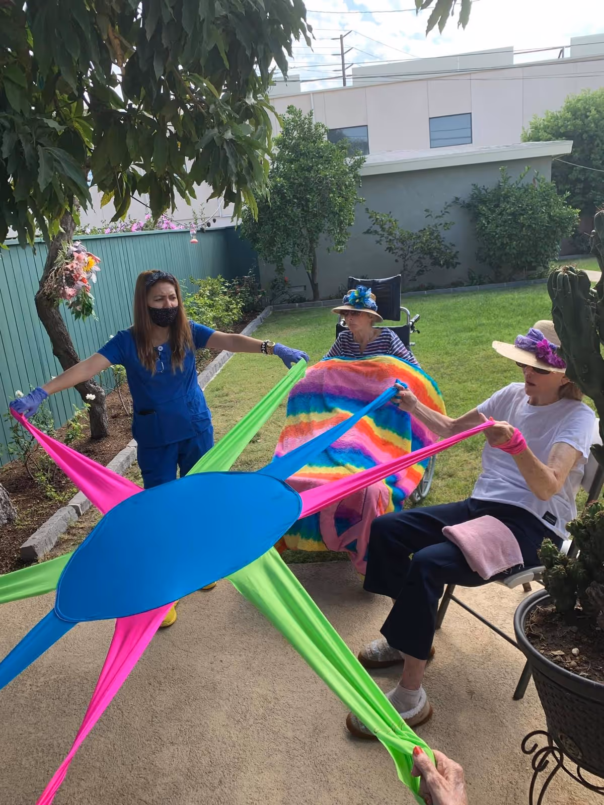 Three elderly women and a caregiver outdoors in a garden area at Palm Gardens Senior Wellness Home. One woman is in a wheelchair covered with a colorful blanket, another woman is seated on a chair wearing a hat, and the caregiver is standing. They are all holding onto a large, colorful stretchy fabric exercise tool. The setting is a green lawn with trees and plants around.